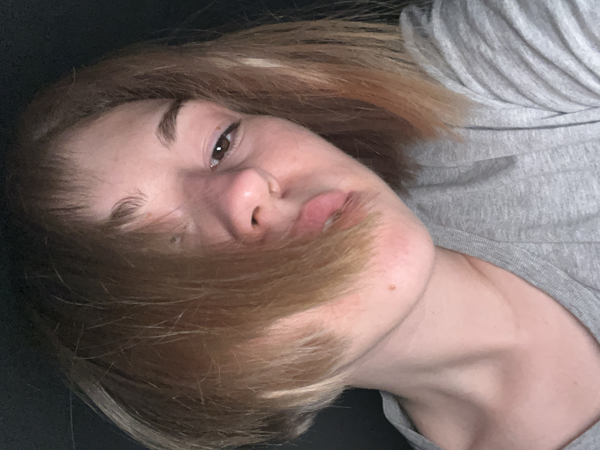 A close-up, angled, and somewhat blurry image shows a woman's face. She has light brown hair with blonde highlights. Her skin tone is fair, with some redness around the nose. She has brown eyes, defined eyebrows, and appears to be wearing a grey t-shirt. Part of her hair is falling across her face. The background is dark and out of focus. There is no evidence to suggest a location in the city of Boreti, Montenegro. The photo appears to be taken indoors in low light.