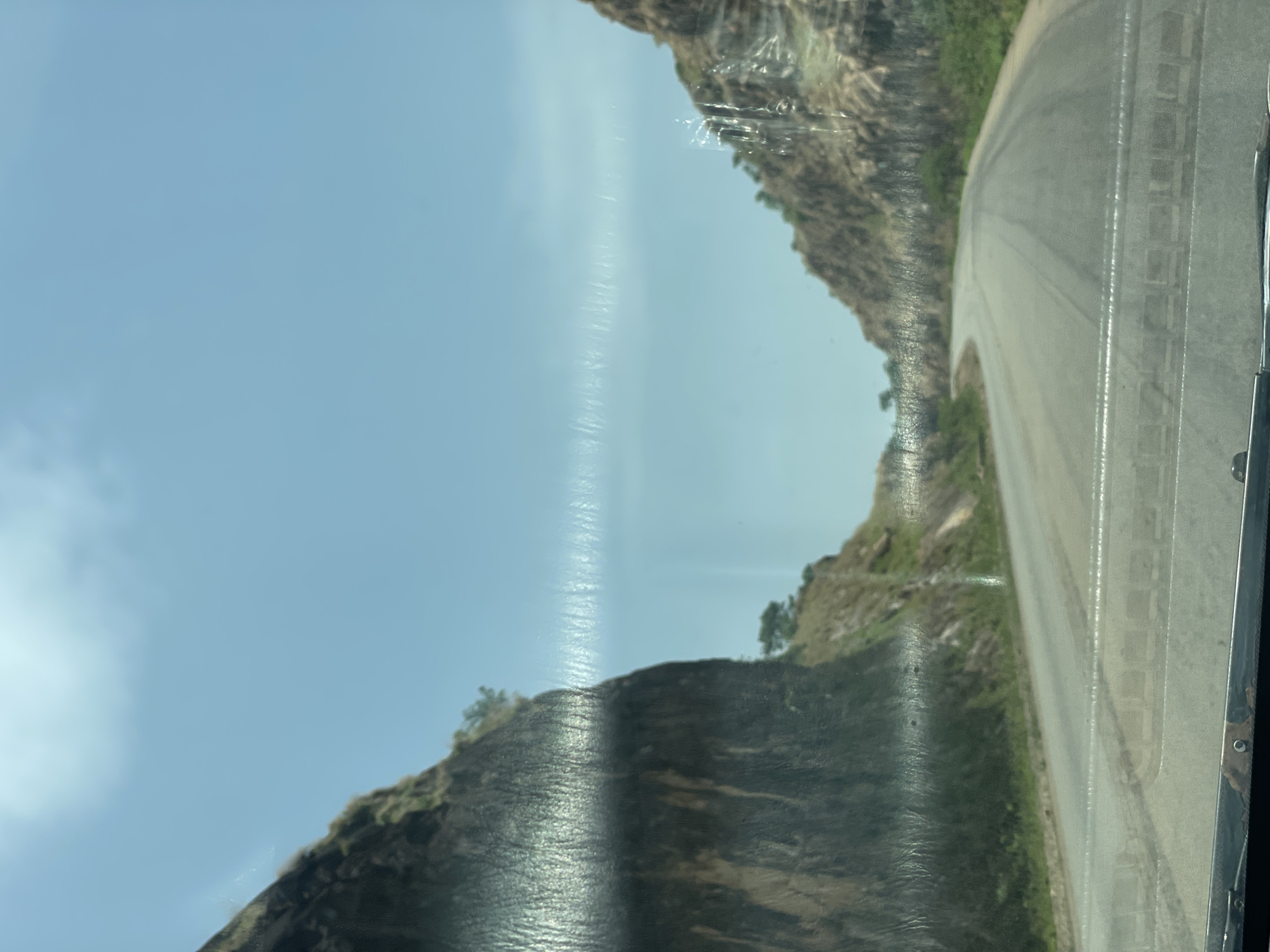The image, seemingly taken from inside a moving vehicle, shows a road winding through a rocky, mountainous area. The sky is pale blue with some cloud cover. The road surface is gray and shows signs of wear. On either side of the road, steep rocky slopes rise up. There is sparse vegetation on the slopes, including some trees. The vehicle's windshield is slightly dirty and shows some streaks or reflections. Based on the location provided, this scene is likely a rural area in Vom, Nigeria, known for its rocky terrain. No people are visible in the image.