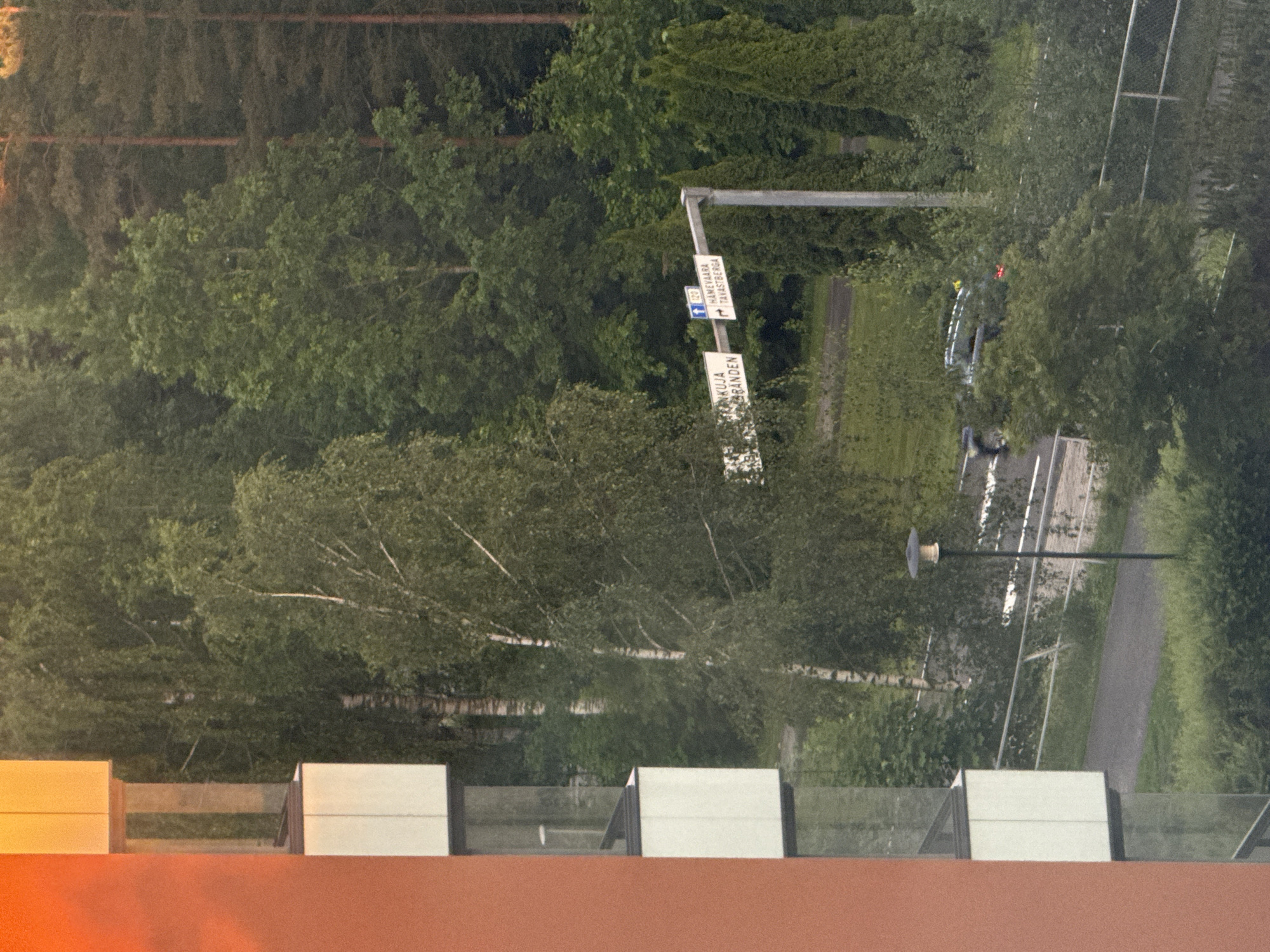 The image shows a view from a building, looking out towards a tree-lined area and a road. On the left side of the image is the exterior of a building with orange and white sections, featuring balconies with glass railings. In the background, there is a dense forest with various types of trees, including birch and pine. A road runs through the scene, with a white line marking its edge. A street lamp is visible to the right of the road, and a white road sign with blue and white text is mounted on a pole. The sign appears to indicate directions and possibly distances. A dark-colored car is seen driving on the road. The overall environment suggests a suburban or semi-rural setting.