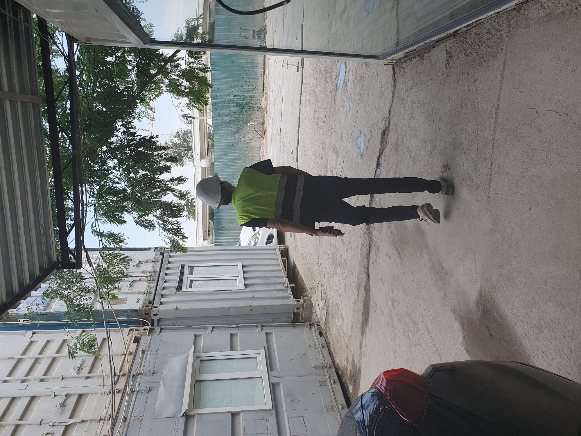 In Tashkent, Uzbekistan, a person wearing a hard hat and a high-visibility vest is walking away from the camera on a concrete surface. They are dressed in dark trousers and tan shoes. Behind them, to the left, are stacked shipping containers with windows. To the right, a large glass-fronted building is partially visible. In the background, trees with green foliage are visible, along with a light-colored fence and a construction crane. A black car is parked on the left side of the image. The overall setting appears to be an outdoor industrial or construction area.