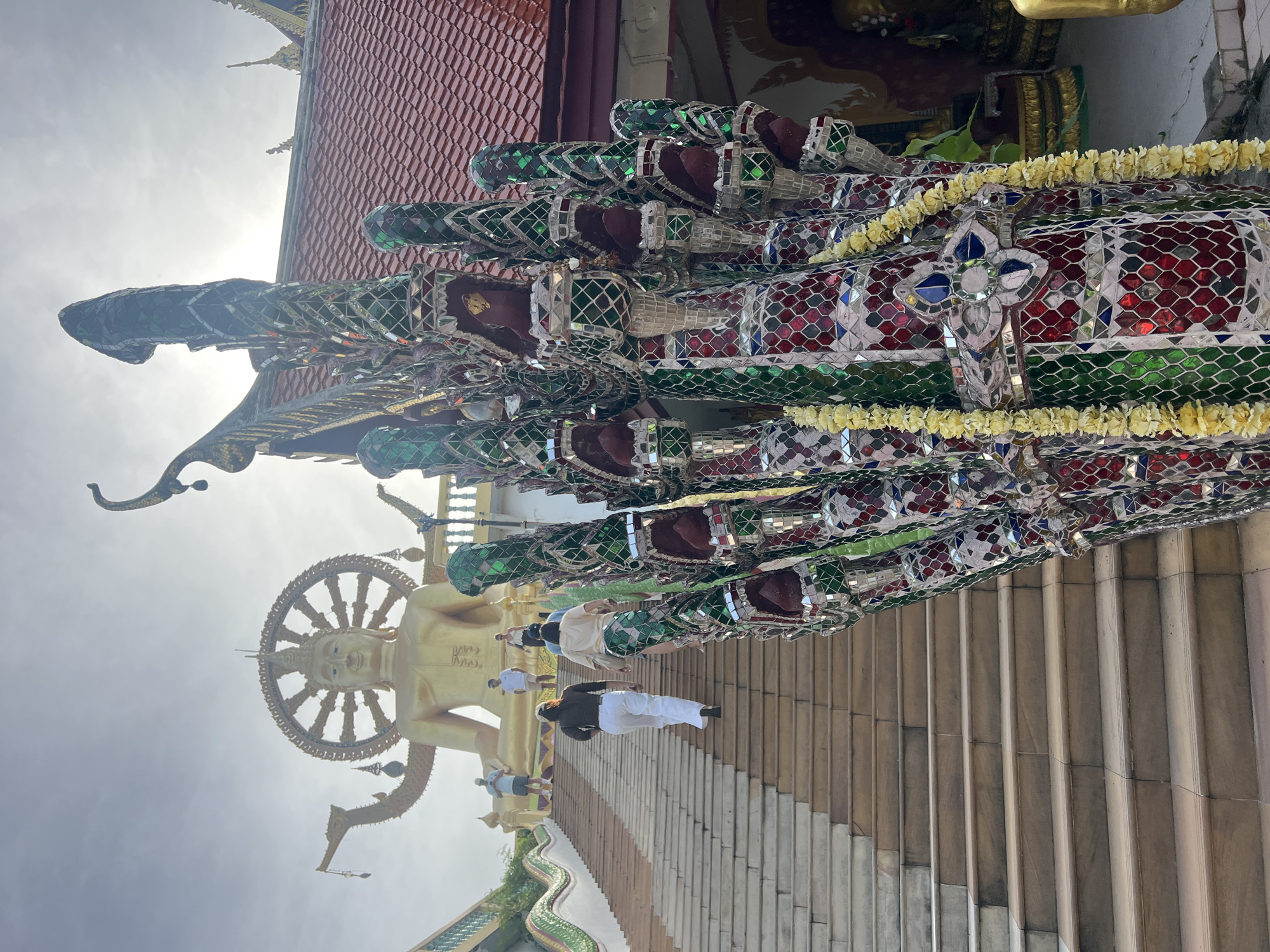 In Baan Plai Laem, Thailand, a wide, stone staircase ascends towards a colossal golden Buddha statue. The Buddha is seated, with a Wheel of Dharma behind it, and its arms are raised in a gesture of blessing. Several people are walking up the stairs, some in casual attire, others in more traditional clothing. In the foreground, a decorative, multi-headed serpent-like sculpture, adorned with colorful mosaic tiles and yellow flower garlands, partially obstructs the view of the stairs. The sky above is bright, suggesting it is daytime, with sunlight filtering through the clouds. The architecture of the temple complex, with its red-tiled roofs, is visible in the background.