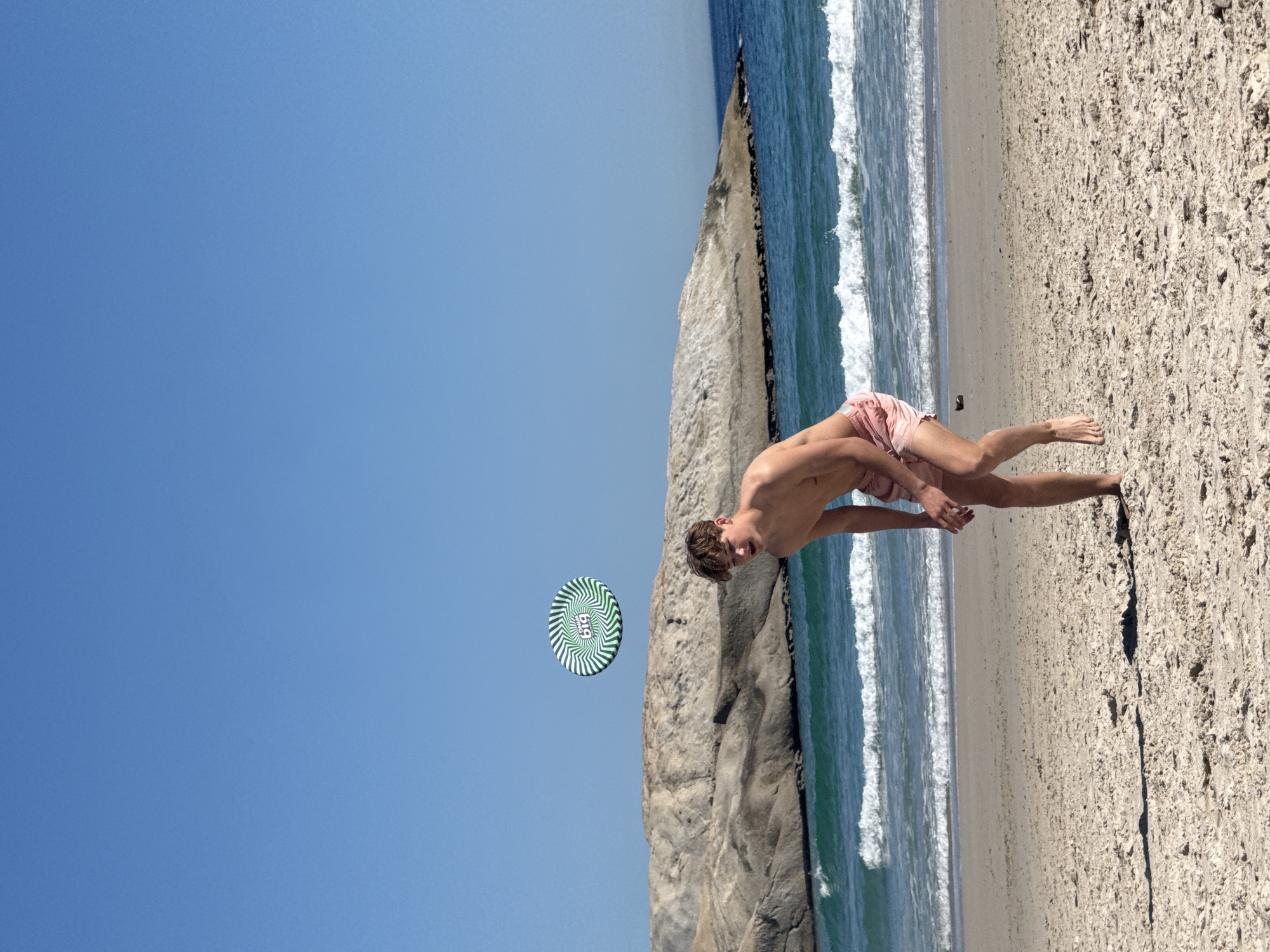 The image depicts a dynamic beach scene in Hout Bay, South Africa, under a bright, clear blue sky. A young male, shirtless and wearing pink swim shorts, is captured in motion on the sandy beach. He leans forward with arms extended, suggesting he is either throwing or catching a frisbee, displaying a focused expression.
In mid-air to the left of the person, a frisbee with a distinctive green and white spiral pattern is visible. The words "Big BIG" are printed in the center of the frisbee.
The background features a light-colored sandy beach extending towards the right, meeting the turquoise-blue ocean. White waves break gently along the shoreline. To the left, a large, light-colored rocky outcrop or cliff face rises from the land, adding rugged texture to the coastal environment. The bright sunlight suggests it is midday or early afternoon, creating a vibrant and active atmosphere.