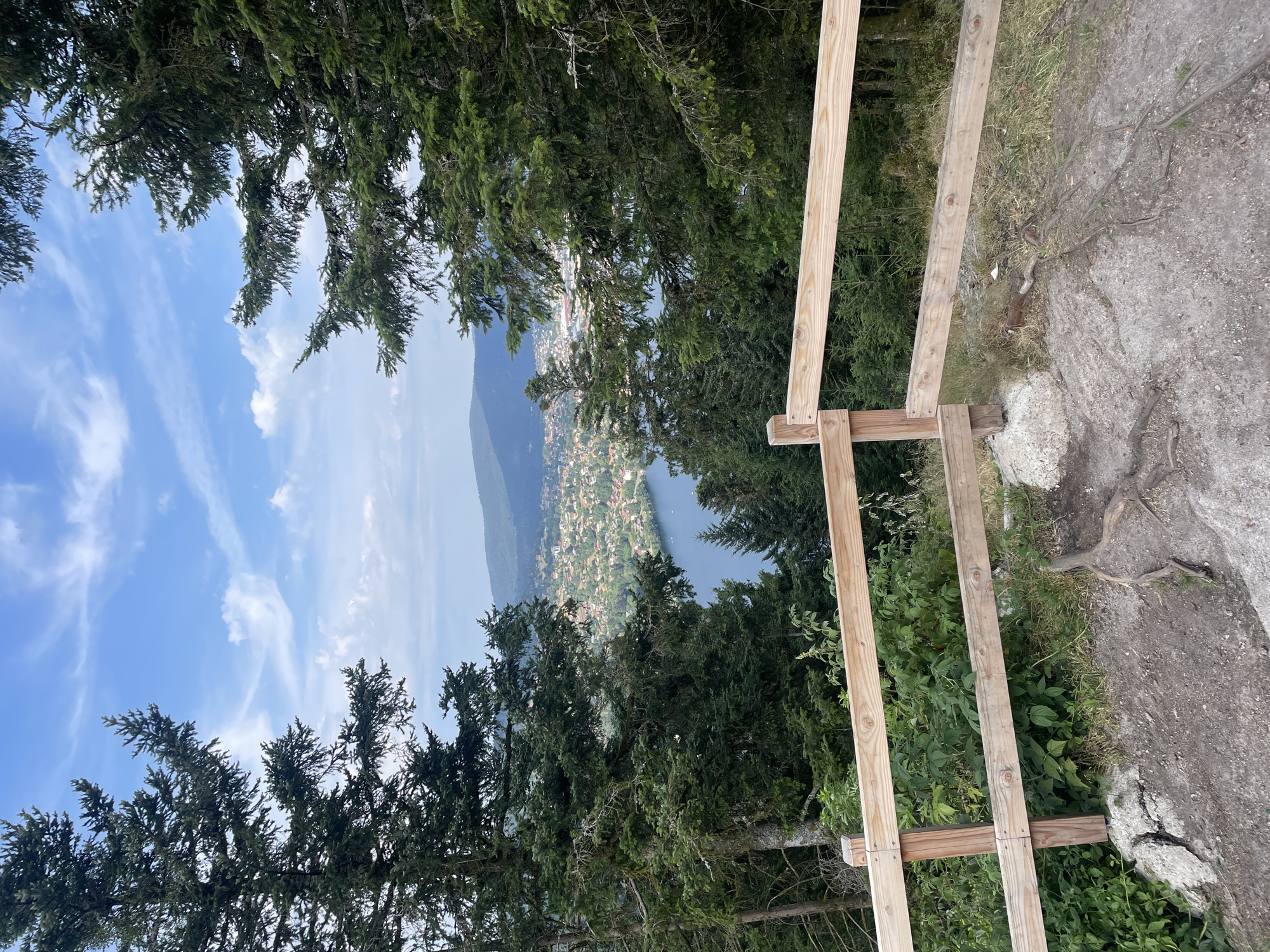 A scenic vista overlooks the city of Gérardmer, France, and its eponymous lake. The foreground features a rustic wooden fence, with two horizontal planks supported by vertical posts, set against a backdrop of lush green foliage and exposed tree roots on the rocky ground. Beyond the fence, dense evergreen trees frame a panoramic view. The mid-ground reveals the sprawling city of Gérardmer, with its cluster of buildings and red-tiled roofs, situated along the serene blue expanse of the lake. In the distance, rolling green hills and mountains rise under a bright blue sky dotted with scattered white clouds.