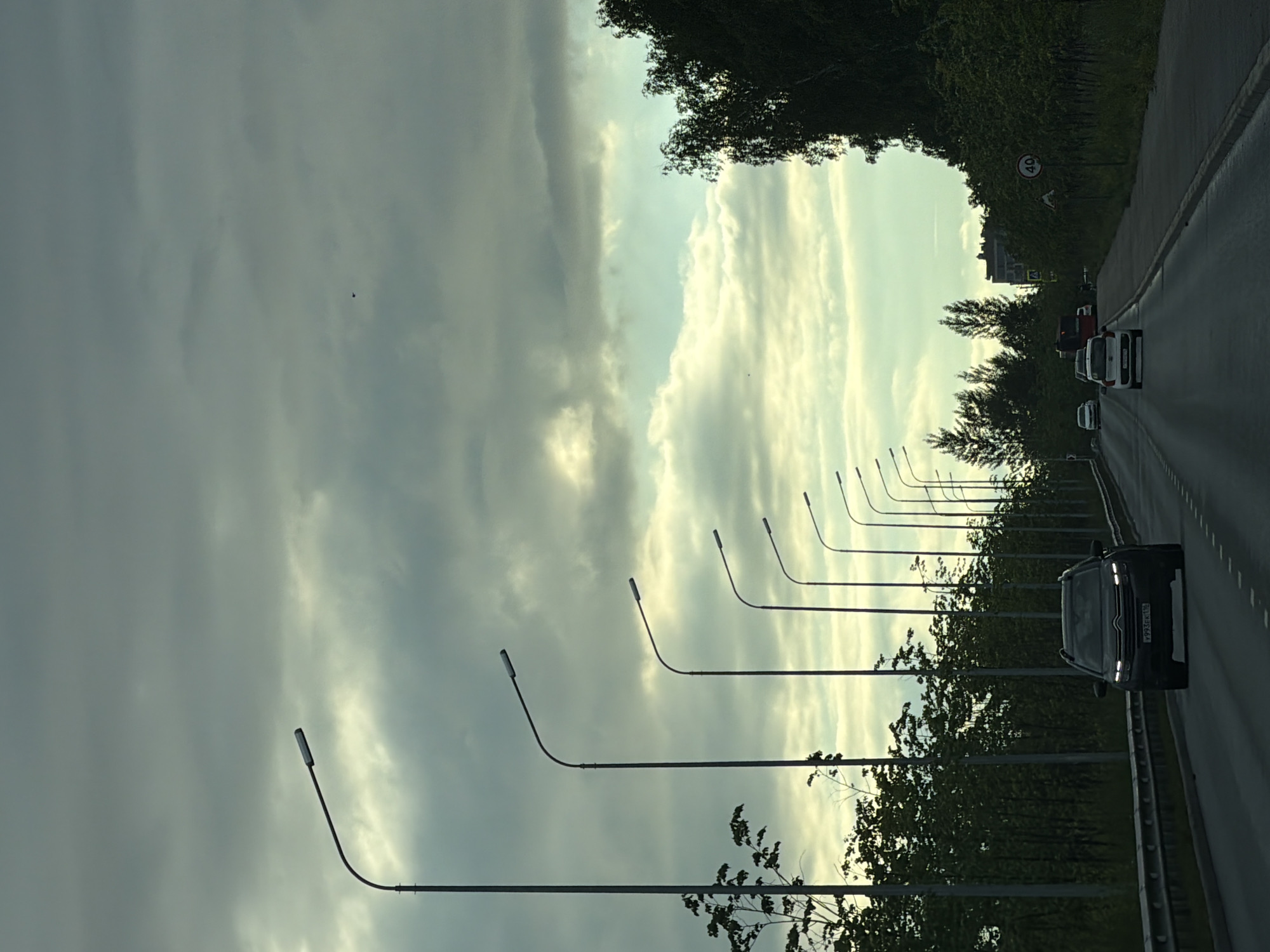 The image shows a road with a line of streetlights on the left side. The road curves to the right and appears to be in a somewhat rural or suburban setting, with trees lining the side of the road. Several cars are visible on the road, traveling in the same direction as the camera. The sky is cloudy, with patches of lighter clouds suggesting it might be late afternoon or early evening. The overall mood is calm and ordinary, depicting a typical drive.
The primary objects in the scene are the streetlights, which are evenly spaced along the road and are all turned off. There are also several vehicles, including a black SUV in the foreground that appears to be leading the pack of cars. Further down the road, other cars are visible. The road itself is paved and has lane markings.
The setting appears to be a road surrounded by greenery. The trees are dense and dark, contrasting with the lighter sky. The clouds are varied in texture and density, some appearing wispy and others more solid.
There is a speed limit sign visible on the side of the road with the number "40" on it. The black SUV in the foreground has a license plate with text, but it is difficult to read clearly.
The time of day is not definitively clear but the lighting suggests it is not midday. The weather is overcast or partly cloudy. No people are clearly discernible as subjects. The scene depicts travel on a road.
