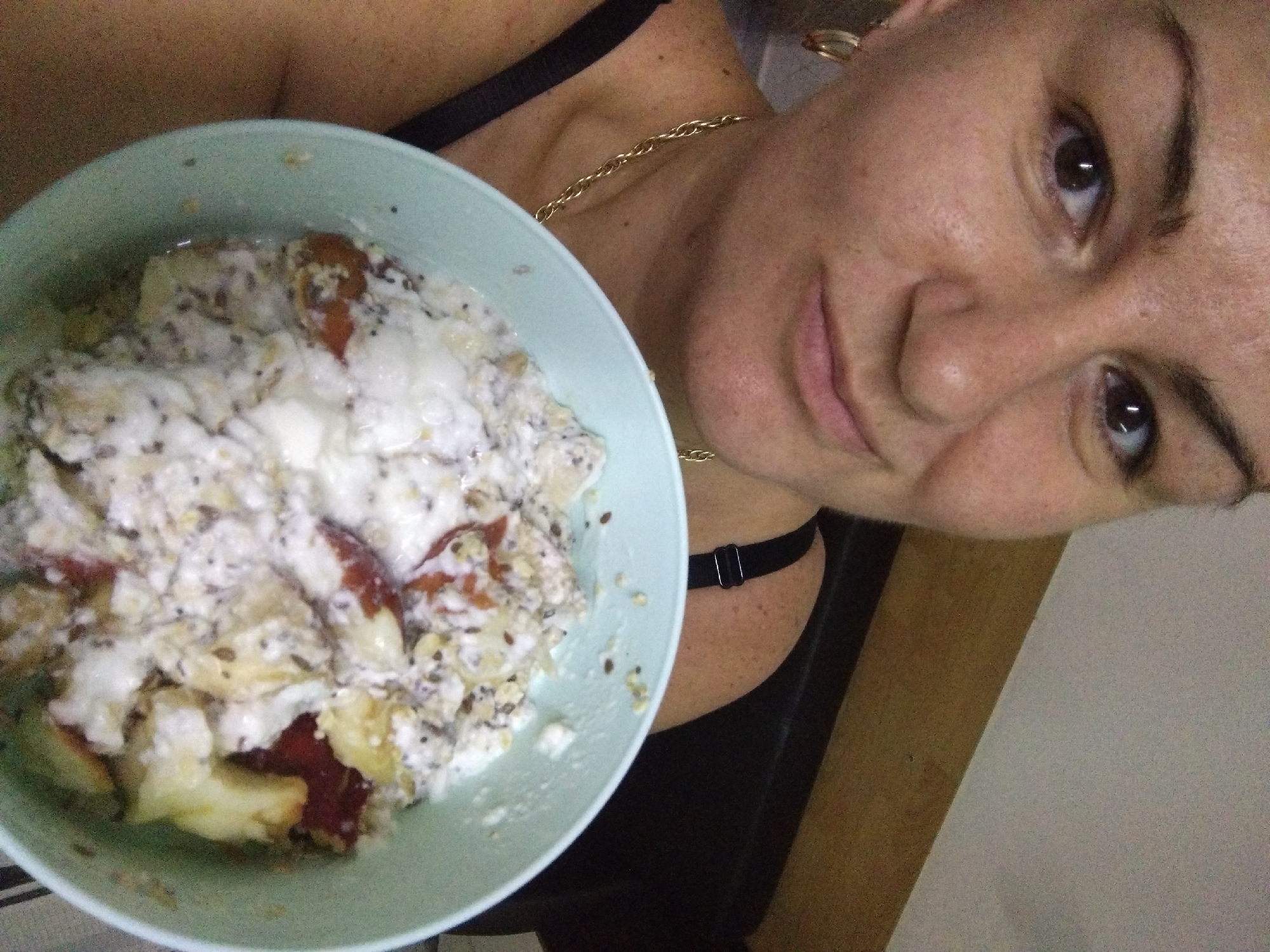 A woman holds a light blue bowl filled with food, likely oatmeal or yogurt with fruit and seeds. The bowl is positioned on the left side of the frame, close to the camera. The food in the bowl appears to contain chopped apples, possibly other fruits, oatmeal, yogurt or a similar dairy product, and seeds like chia or flax. The woman is facing the camera, with her face occupying the upper right portion of the image. She has fair skin, brown eyes, and her hair appears to be short. She is wearing a black tank top and a gold chain necklace. An earring is visible in her left ear. The background is out of focus, suggesting an indoor setting. There is a light-colored wall behind her and a darker wooden panel or surface on the right side of the frame. There's no evidence suggesting the location is Stara Zagora, Bulgaria.