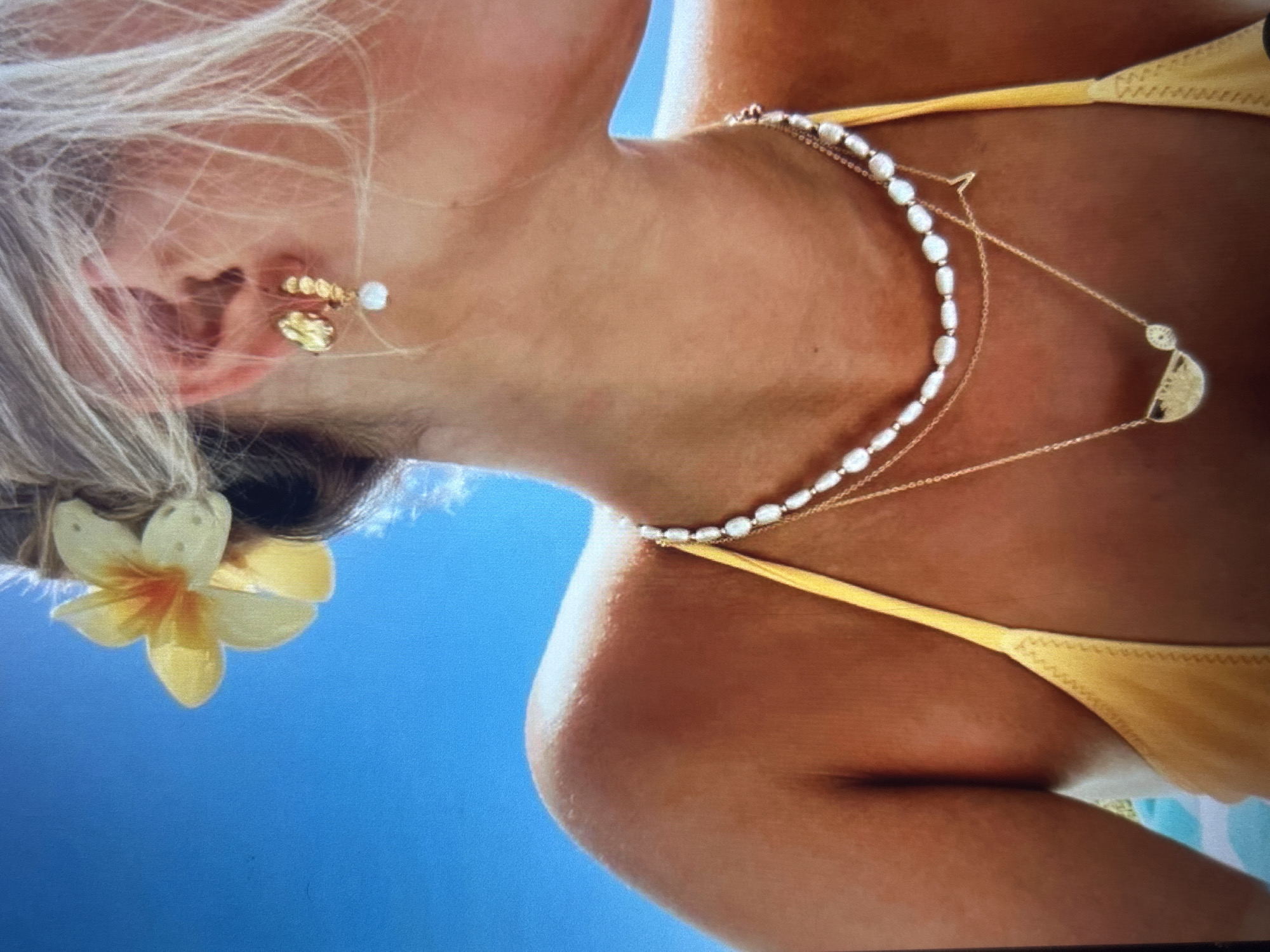 The image shows a close-up view of a person wearing jewelry and a yellow bikini top against a bright blue sky. A yellow flower is placed in the person's hair. The person is also wearing multiple necklaces: one with pearls, and two delicate gold chains, one with a small "V" pendant and the other with a crescent-shaped pendant. There is also a gold earring in the visible ear. The image does not contain any information that would allow for a determination of its location in the city of Knezha, Bulgaria.