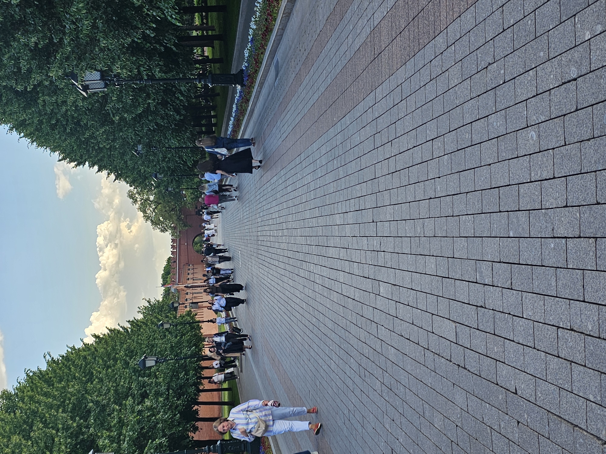 A wide, paved pedestrian pathway stretches into the distance, flanked on both sides by rows of tall, leafy green trees. The path is constructed from gray rectangular paving stones, featuring a central stripe of reddish-brown blocks. To the right, a flower bed with various colorful blooms runs alongside the path. On the left side, behind the trees, a distinctive red brick wall, characteristic of a historical fortress or city wall with battlements, is visible, culminating in an arched entrance further down the path.
Numerous people are seen walking along the pathway in various directions. In the foreground, a woman wearing a light-colored plaid shirt and light-blue pants, carrying a beige bag, walks towards the camera. Other individuals and small groups are scattered along the length of the path, engaged in casual strolling or sightseeing. Ornate black streetlights are interspersed among the trees.
The scene depicts a pleasant daytime setting, under a partly cloudy sky with bright white clouds. The overall impression is one of leisure and public access to a potentially historic or scenic area. No visible text is discernible in the image.