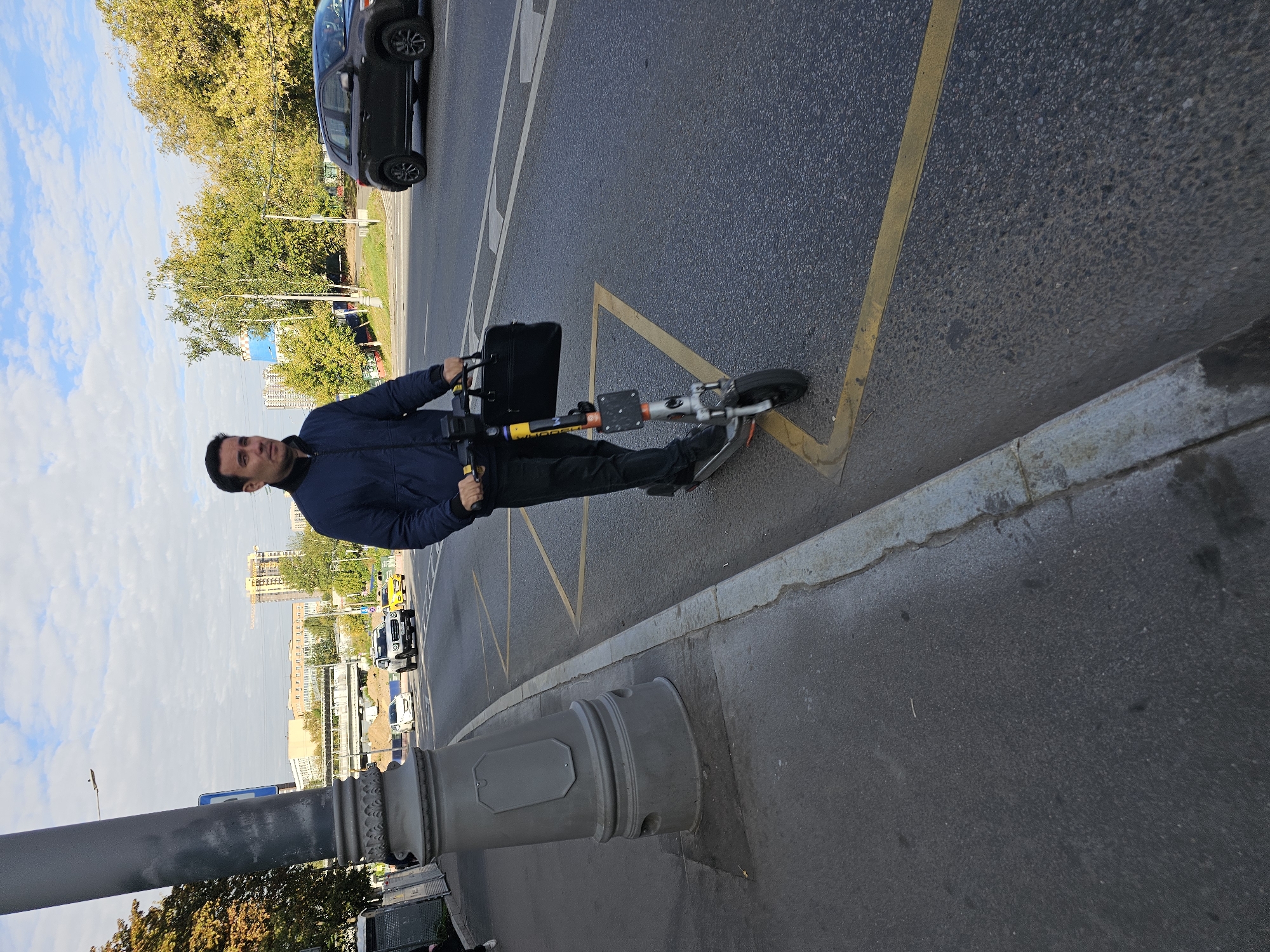 A man is riding an electric scooter on the side of a road in Moscow, Russia. He is dressed in a dark blue jacket and black pants and is holding a black briefcase in his right hand. The scooter is silver and orange. In the background, there are several cars on the road, trees, and buildings. The sky is cloudy. The man is positioned on the right side of the image, riding away from the camera. The scooter is on the asphalt road, next to a sidewalk and a large grey lamppost. The road has yellow markings indicating a bus stop or no-parking zone. The environment appears to be an urban setting with a mix of greenery and infrastructure.