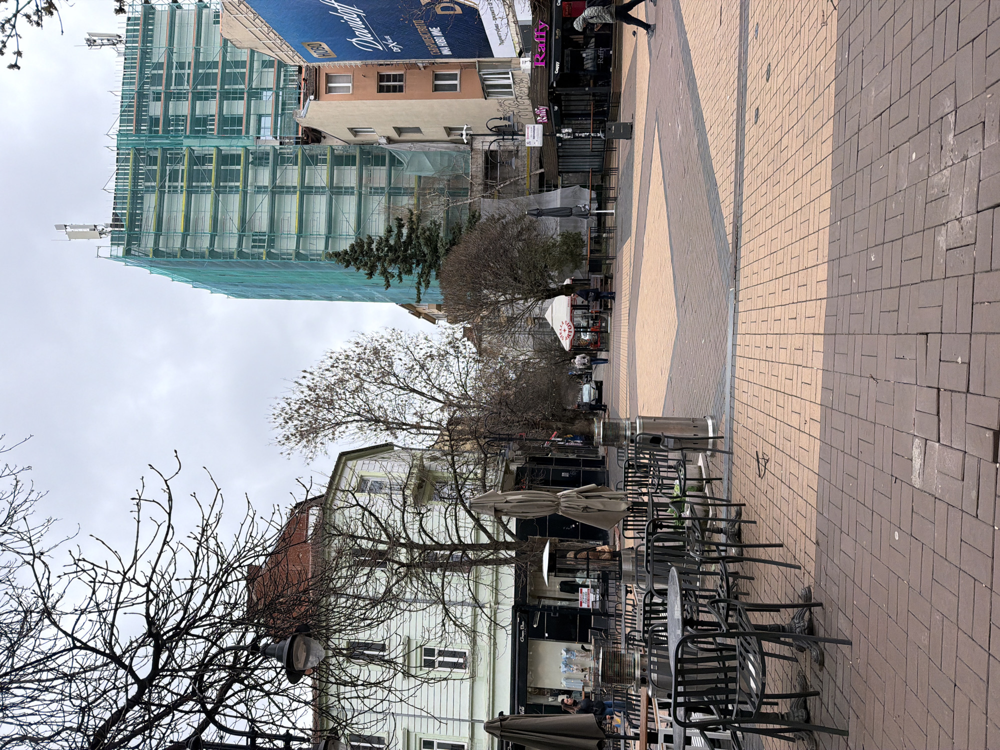 The image shows a street scene in Sofia, Bulgaria. The view is looking down a pedestrianized street paved with interlocking bricks. Tables and chairs from an outdoor cafe line the right side of the street. Buildings are visible on either side. On the left, a tall building is under construction with scaffolding covered in green netting. A large blue sign advertising "Davidoff" is visible at the top of one of the buildings. The sky is overcast. Bare trees and a street lamp appear in the foreground. People are walking on the right-hand side of the street.