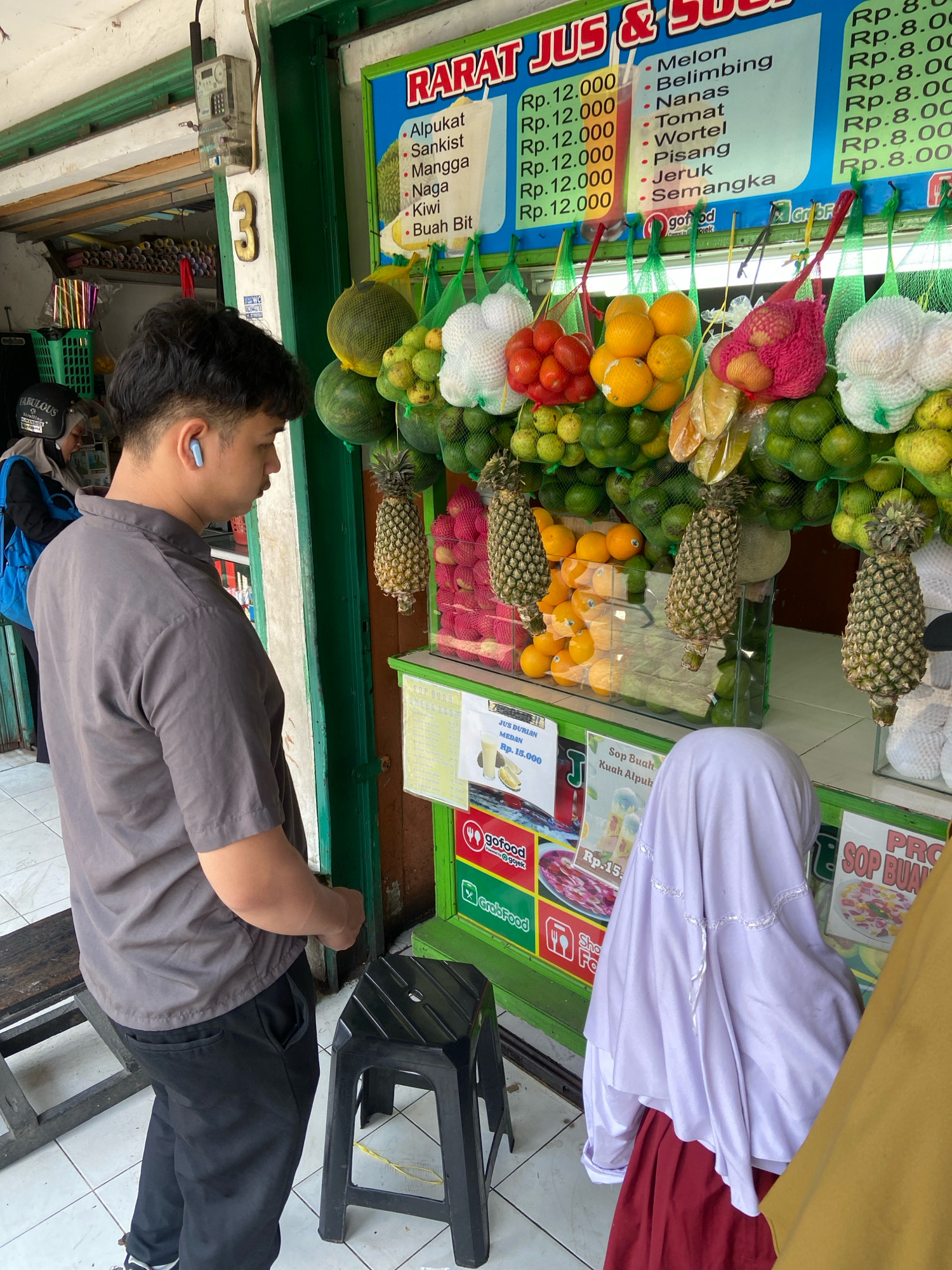 In North Jakarta, Indonesia, a young man wearing a gray collared shirt and black pants stands at a juice stand. He has dark hair and is wearing a blue wireless earbud. The juice stand displays a variety of fresh fruits such as watermelons, oranges, pineapples, and coconuts. A price list with juice options like melon, pineapple, and tomato is visible. In the background, a woman in a headscarf and a blue backpack is seen. The scene appears to be outdoors, possibly a street or market area, with a tiled floor and a green painted structure. The overall atmosphere suggests a bustling urban environment, common in areas like North Jakarta.