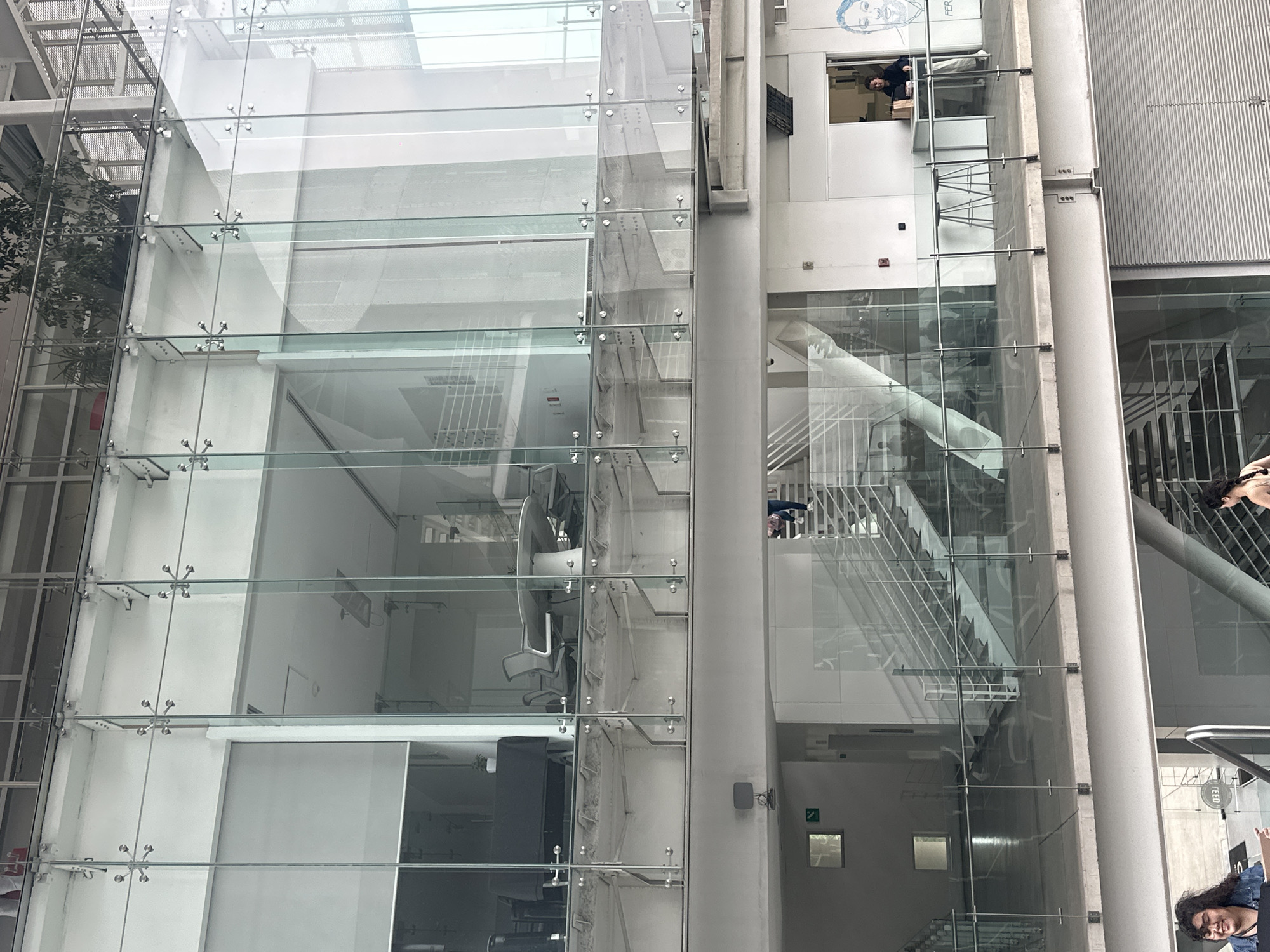 The image shows the interior of a modern building in Mexico City, Mexico. The architecture features a large glass facade with steel supports, offering a view into the multi-story structure.
Inside, there are open office spaces visible through the glass, with office furniture like chairs and desks. Staircases are prominent, constructed with glass and metal, providing access to different levels. There are people visible on different floors of the building.
The building has an overall clean and contemporary aesthetic, with a neutral color palette of white and grey. The presence of people indicates that the building is in use.