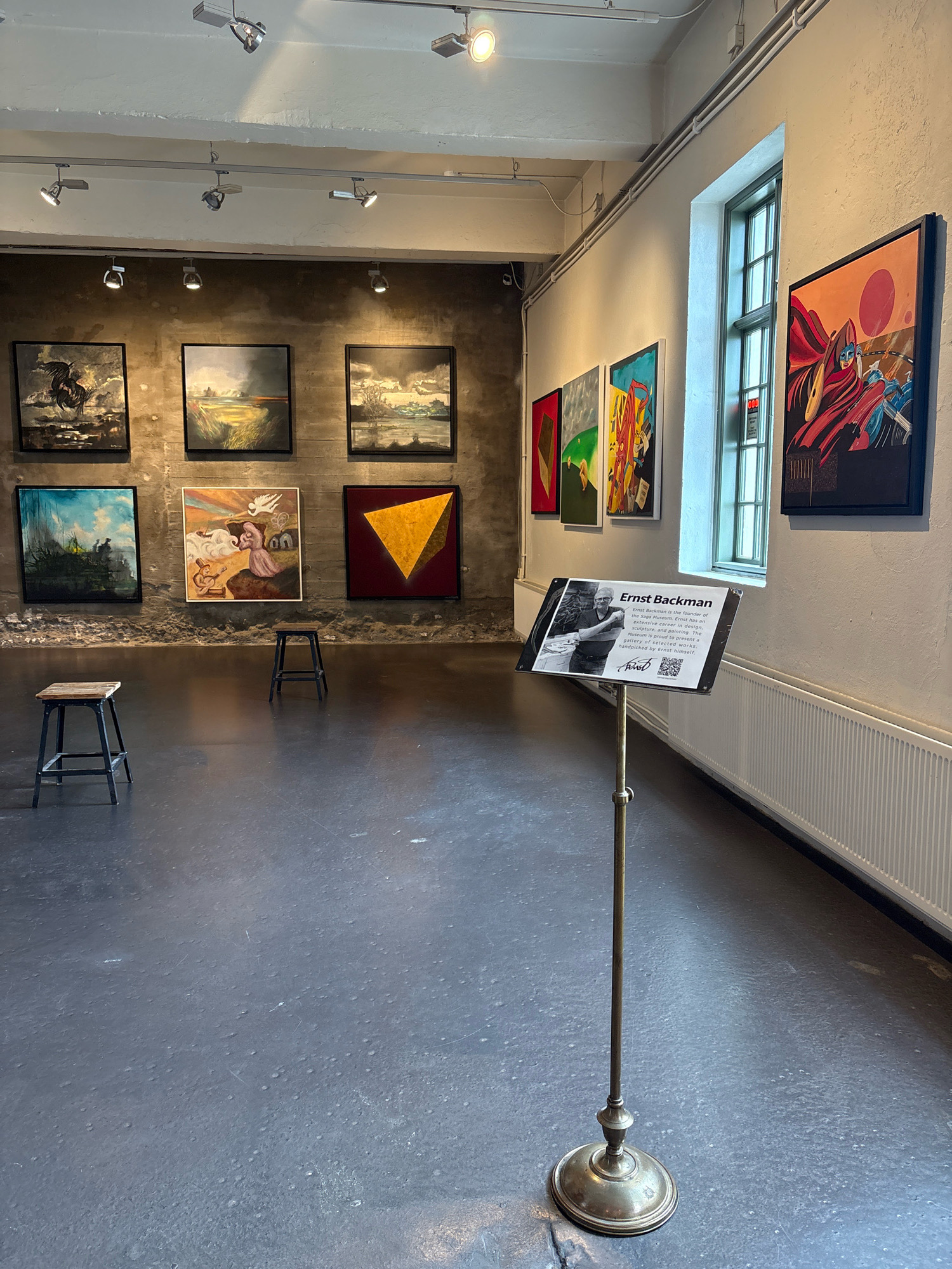 The image depicts the interior of an art gallery, likely in Reykjavik, Iceland, given the context provided. The scene is brightly lit by overhead track lighting. The walls are a rough, concrete texture on the left and a plain white on the right. Several framed paintings are displayed on the walls, featuring a variety of styles from landscapes to abstract and figurative works. In the foreground, a brass sign on a stand provides information about "Ernst Backman," identifying him as the founder of the Saga Museum and a collector of art. Two simple wooden stools are placed in front of some of the paintings, suggesting seating for viewers. A large window with multiple panes is visible on the right side of the room, and a white radiator runs along the bottom of the wall beneath it. The floor is a dark, polished surface. The overall impression is of a clean, modern gallery space showcasing a diverse collection of artwork.