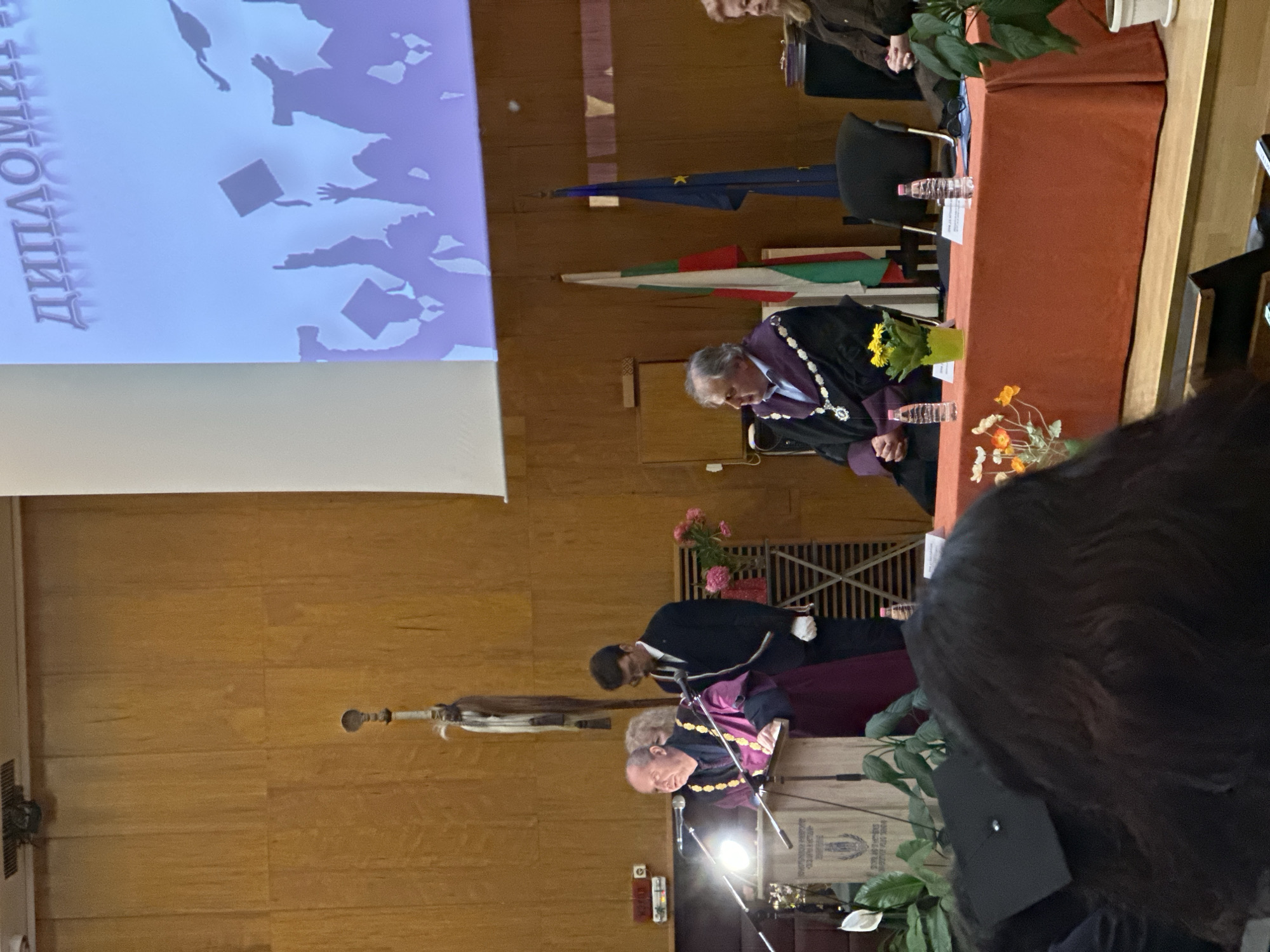 The image shows an indoor event, possibly a graduation ceremony, in Veliko Tarnovo, Bulgaria. A screen displays graphics. The Bulgarian and European Union flags are visible. At least two individuals wearing academic gowns are present, one seated behind a table covered with an orange cloth. Flowers and water bottles are on the table. A second person is bowing to a seated person. The room has wood paneling. A ceremonial mace is visible in the background.