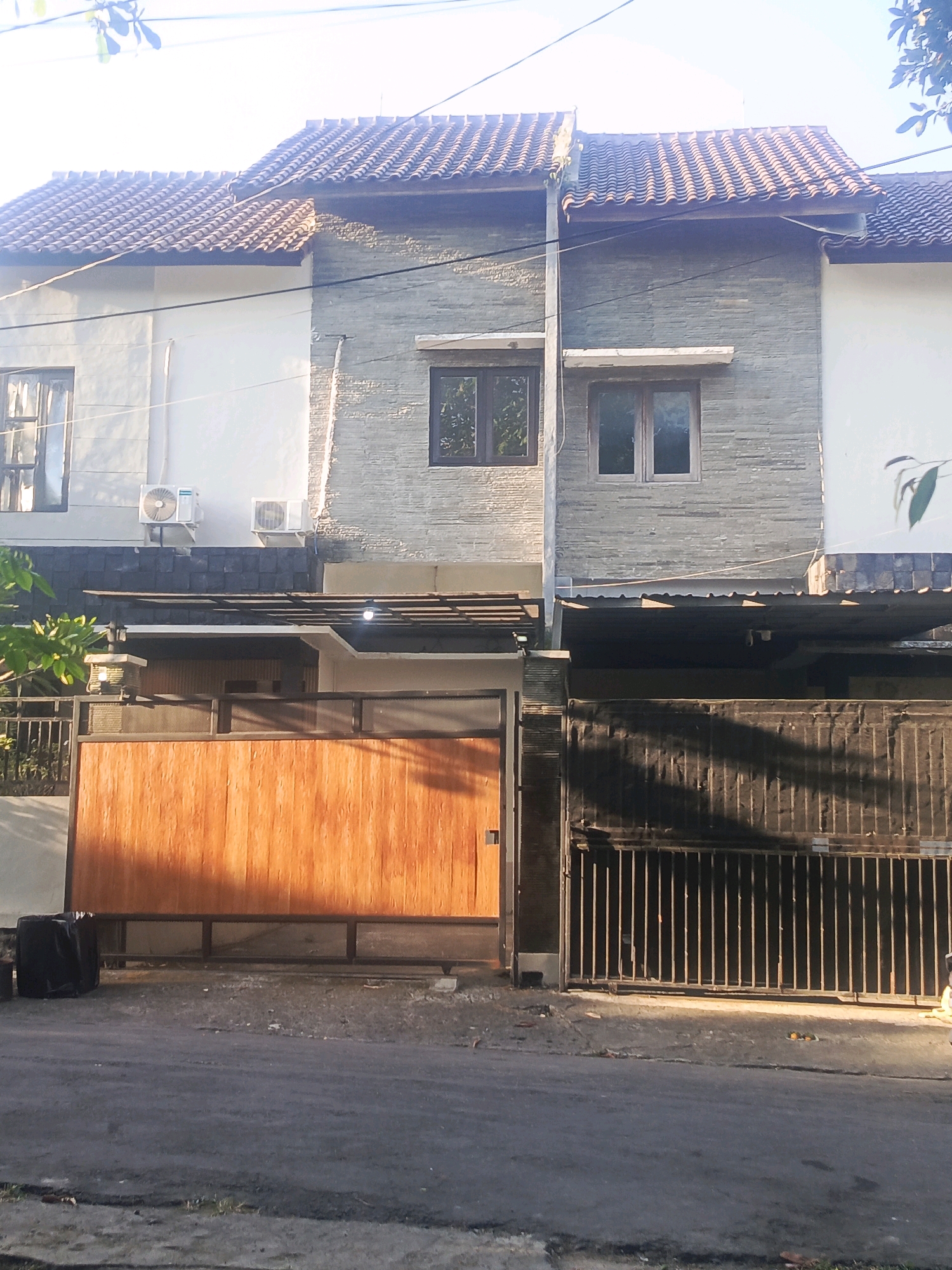 The image shows a row of modern townhouses in North Kuta, Indonesia. The houses have tiled roofs, stone and white stucco facades, and dark metal gates. The property on the left has a large, solid wooden gate, while the property on the right has a black metal bar gate. Two air conditioning units are visible on the exterior wall of the left townhouse. The street in front of the houses is paved and has some debris on the shoulder. Trees and foliage are visible in the background. The lighting suggests it is either early morning or late afternoon, with long shadows cast across the scene.