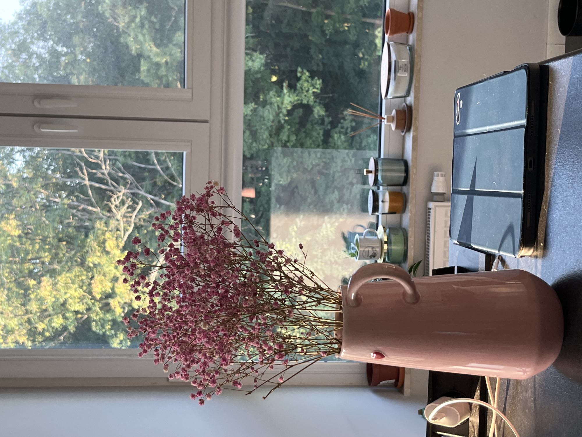 In an indoor scene by a window, a light pink ceramic pitcher filled with a bouquet of small pink flowers is placed on a dark countertop. The pitcher has a handle and a small red heart design. To the left of the pitcher, a white electrical outlet with a charging cable is visible. On the windowsill behind the pitcher, several objects are arranged: a small terracotta pot, a white mug with a design, a jar with a yellow lid, a light blue jar with a wooden lid, a reed diffuser with a wooden base, and a large white jar with a wooden lid. Beyond the objects on the windowsill, a window looks out onto a dense forest with trees, illuminated by soft sunlight. In the foreground on the countertop, a black tablet with a patterned case is positioned to the right of the pitcher.