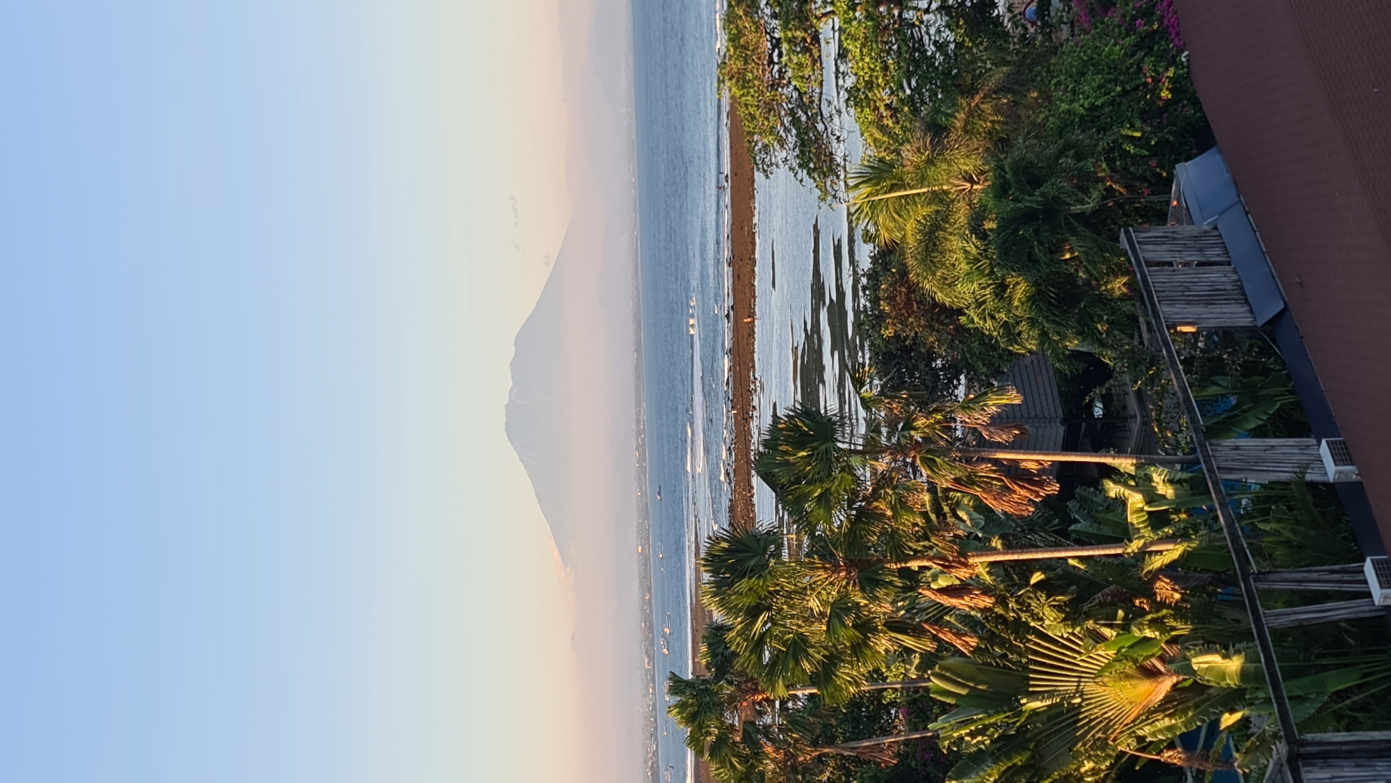 An elevated, vertical, full shot depicts a serene coastal landscape at dawn or dusk. A prominent, mist-shrouded mountain dominates the background, its peak barely visible above a hazy horizon. Below the mountain, a vast expanse of calm, blue ocean stretches across the midground, dotted with several small boats. A narrow strip of coastline, appearing to be a beach or rocky shore, separates the ocean from shallow, tide-pool-like water in the foreground. Lush, tropical vegetation, including palm trees with fronds catching the warm light, fills the lower portion of the frame. A wooden fence and parts of a building with a reddish-brown roof are visible in the extreme foreground, suggesting a vantage point from a hotel or resort. The sky is a soft gradient of pale blue, transitioning to a warmer hue near the horizon. The overall atmosphere is tranquil and picturesque, likely capturing a view of Mount Agung from a location in or near Denpasar, Bali, Indonesia.
