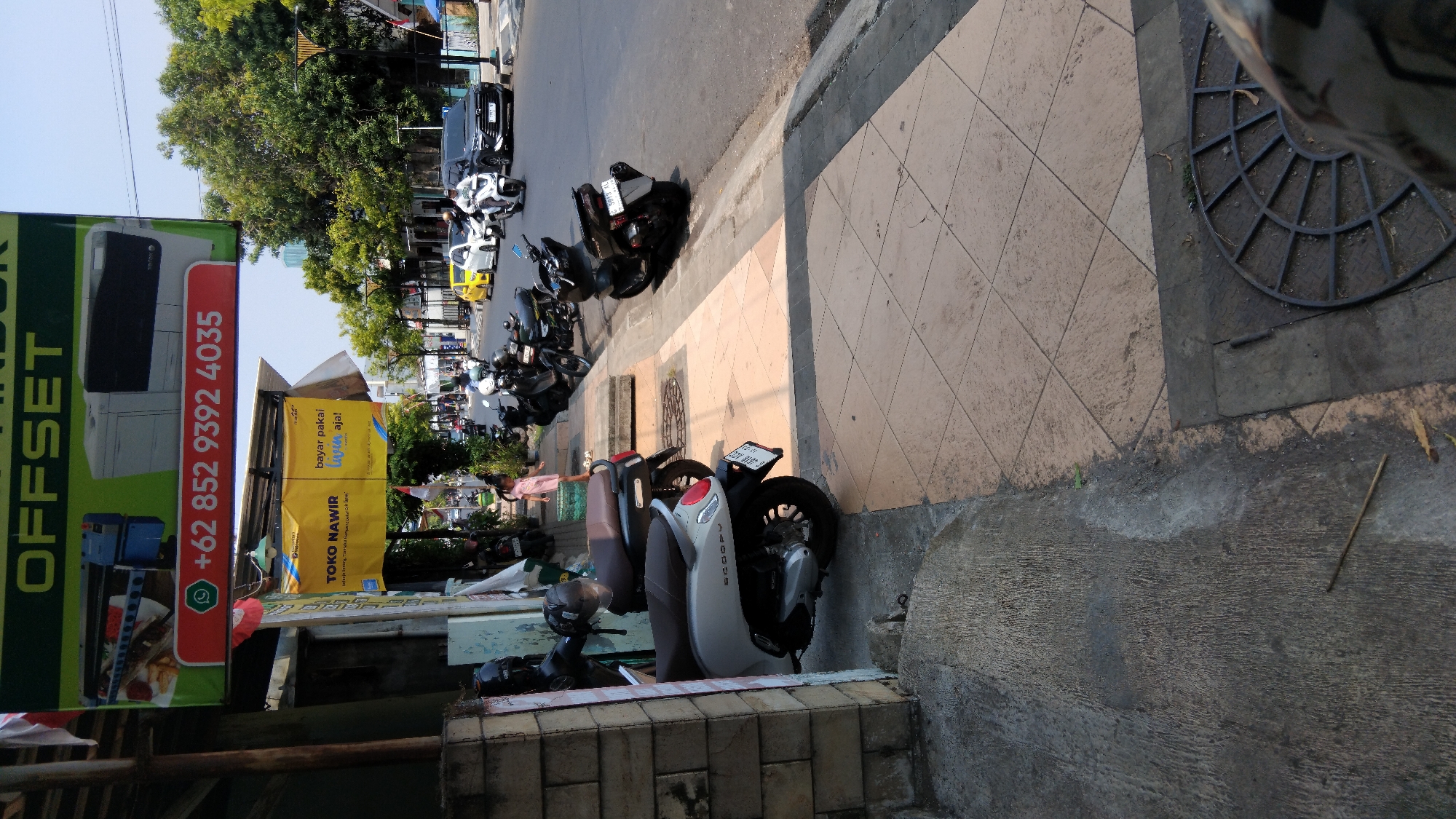 The image depicts a street scene in Batang, Indonesia, with a focus on a sidewalk area and the adjacent road. In the foreground, two scooters are parked on the sidewalk. One is a silver scooter with "SCOOPY" visible on its side, and the other is a darker-colored scooter. A woman in a light pink dress stands on the sidewalk near the scooters, looking towards the road.
Above the sidewalk, a banner is displayed with green and red elements. The green section features the word "OFFSET" prominently, and below it, an image of a printer. The red section contains a phone number: "+62 852 9392 4035", preceded by a WhatsApp icon. Another yellow banner, likely advertising a business, hangs below the main banner with text that appears to include "TOKO NAWIR" and "bayar pakai" followed by "Luwis aja!".
The road in the background shows traffic, including several motorcycles and cars. A yellow car is visible among the vehicles. Trees line the side of the road, and buildings are present in the distance. The sidewalk itself is paved with square tiles, some of which are lighter in color and arranged in a diamond pattern. A circular metal grate is also visible on the sidewalk. The overall environment suggests a commercial or semi-commercial area within a city.
