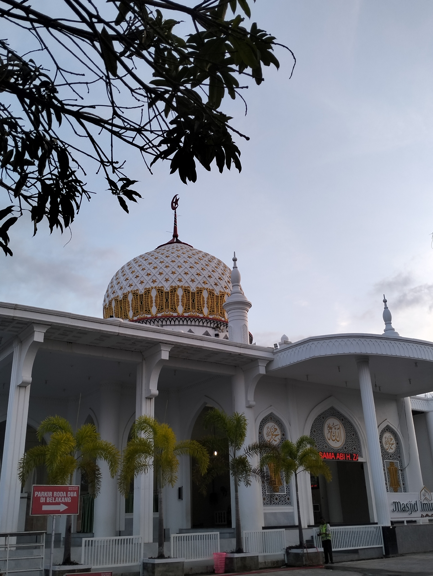 The image shows the exterior of a mosque in Blangmetimu, Indonesia. The mosque features a large, ornate dome decorated with white and gold stars and intricate gold patterns. Two smaller spires are visible on the roofline. The building is predominantly white, with arched windows and columns supporting a covered walkway. In front of the mosque, there are several palm trees and a red sign with white text indicating "PARKIR RODA DUA DI BELAKANG" and an arrow pointing left, suggesting parking for motorcycles is located behind the building. The sky is overcast, and a silhouette of tree branches extends from the top left corner of the frame. A person in a high-visibility vest is standing near the entrance of the mosque. The overall scene depicts a religious building in a tropical or sub-tropical environment.