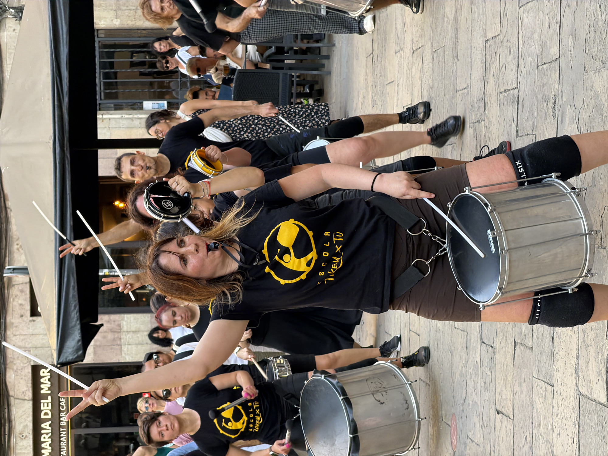In Barcelona, Spain, a group of people, primarily women, are participating in an outdoor parade or performance. The foreground is dominated by a woman in the center, wearing a black t-shirt with a yellow logo that reads "ESCOLA 70KEM X TU," brown shorts, and black knee pads. She is holding drumsticks and has a whistle in her mouth, appearing to be playing a drum that is suspended in front of her. To her left, another woman in a similar black t-shirt is also holding drumsticks and appears to be playing a drum. Several other individuals are visible in the background, some holding drumsticks and others holding small tambourines. They are walking on a cobblestone street lined with buildings, one of which has a sign that reads "MARIA DEL MAR." The event appears to be a festive street performance.