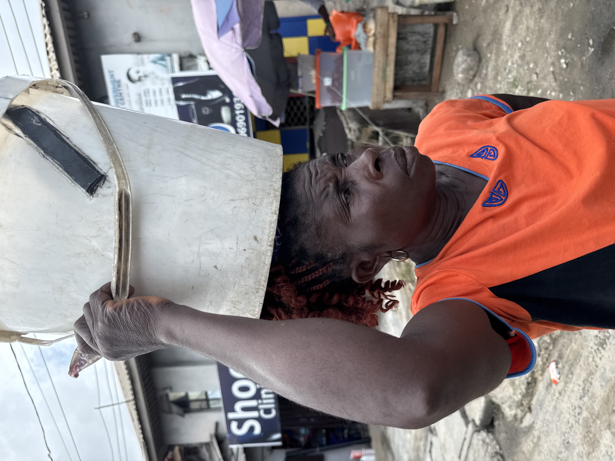A middle-aged, dark-skinned woman in an orange and black t-shirt is carrying a large, white bucket on her head in Ado, Nigeria. She is looking up, her expression unreadable. Her hair is styled in reddish-brown braids with curly ends. She wears a hoop earring in her left ear. The bucket appears to have a metal handle reinforced with black tape.
The environment suggests a busy, possibly impoverished urban area. A sign reading "Sho Clinic" is partially visible behind her. There is laundry hanging in the background, as well as some stacked plastic containers and a wooden structure. The ground is concrete.