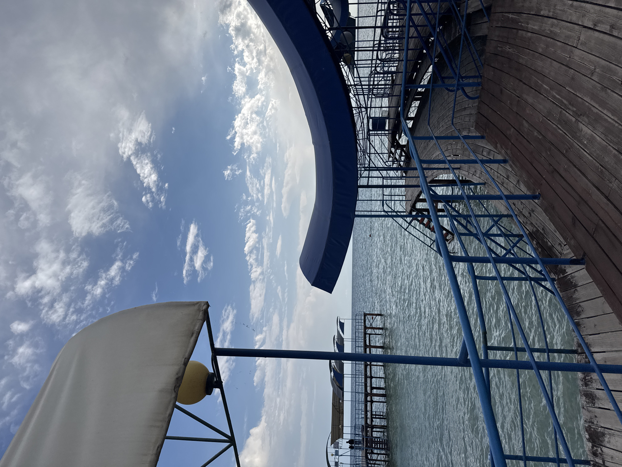 The image depicts a view of a pier or boardwalk extending over a body of water, likely a lake given the location. In the foreground, a wooden walkway with dark planks is visible, bordered by a blue metal railing. The walkway curves to the right, leading towards a structure with a large, curved blue canopy overhead. Stairs with blue railings are integrated into the walkway.
In the midground, a further pier or boardwalk stretches out into the water. This structure also features blue railings and is covered by several smaller blue awnings. Two figures, appearing as silhouettes, are standing on this pier, observing the water. The water itself is calm with ripples on its surface.
The background is dominated by a sky with scattered clouds, some of which are illuminated by sunlight, suggesting it is daytime. The overall impression is a serene, possibly recreational, waterfront scene. The location is specified as Sary-Oy, Kyrgyzstan, which is known for its presence on the shore of Lake Issyk-Kul.