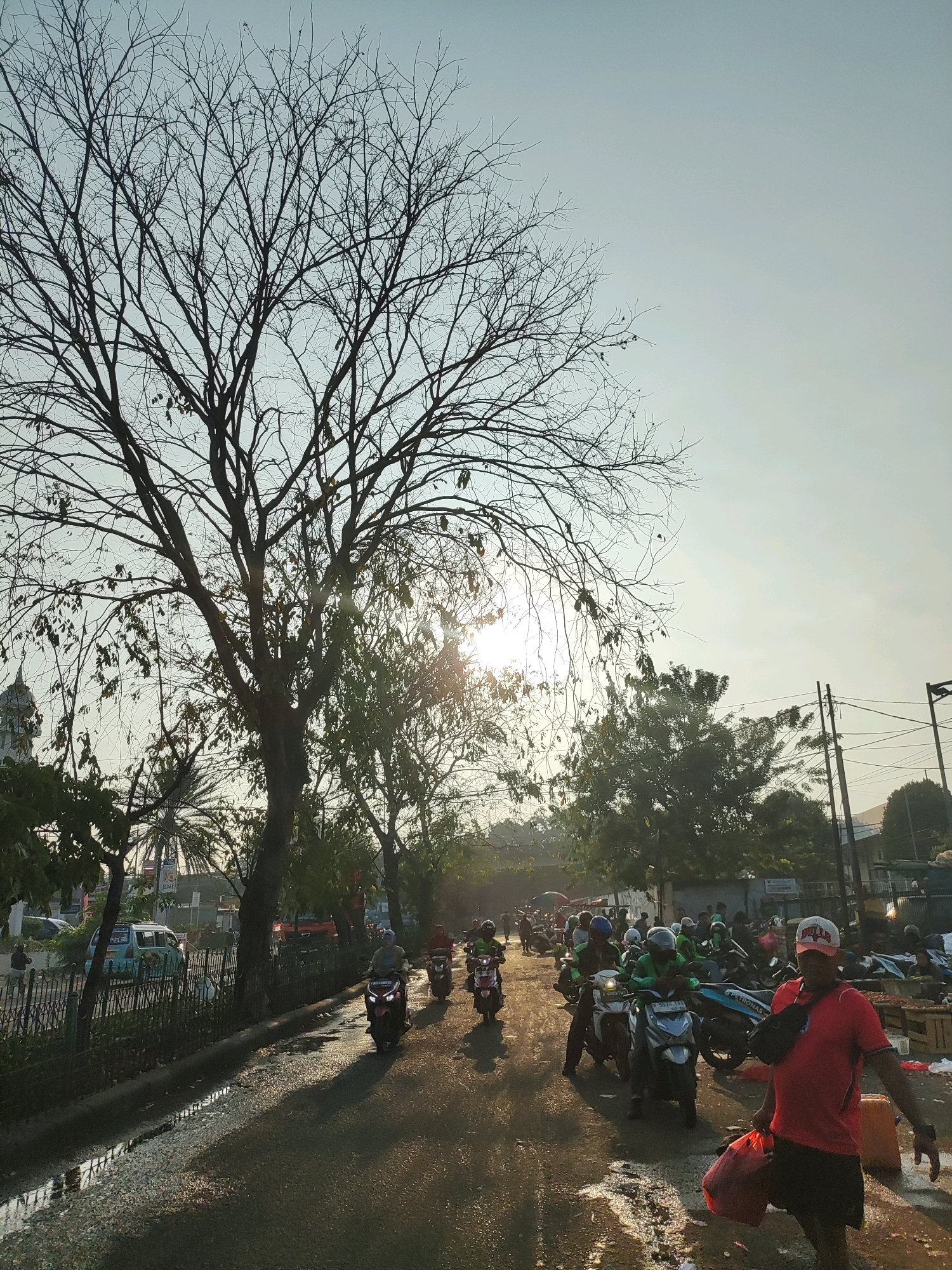 The image captures a bustling street scene in East Jakarta, Indonesia, likely during the early morning or late afternoon due to the low angle of the sun casting long shadows. A large, leafless tree dominates the left side of the frame, its stark branches silhouetted against a pale, hazy sky. Sunlight filters through its branches, creating lens flares.
The street is wet and reflective, suggesting recent rain or a street cleaning operation. A procession of motorcycles, ridden by individuals wearing helmets and casual clothing, moves along the road. Some riders are dressed in green, possibly indicating a delivery service. In the foreground on the right, a man in a red t-shirt and shorts walks towards the viewer, carrying a red bag and a large orange container. He wears a white baseball cap.
Other people are visible in the background, some walking, some on motorcycles. Beyond the road, there is a row of market stalls with produce, and buildings line the street, obscured by trees and power lines. A light blue van is parked on the left side of the street, near a decorative metal fence. The overall atmosphere is one of urban activity and commerce.
