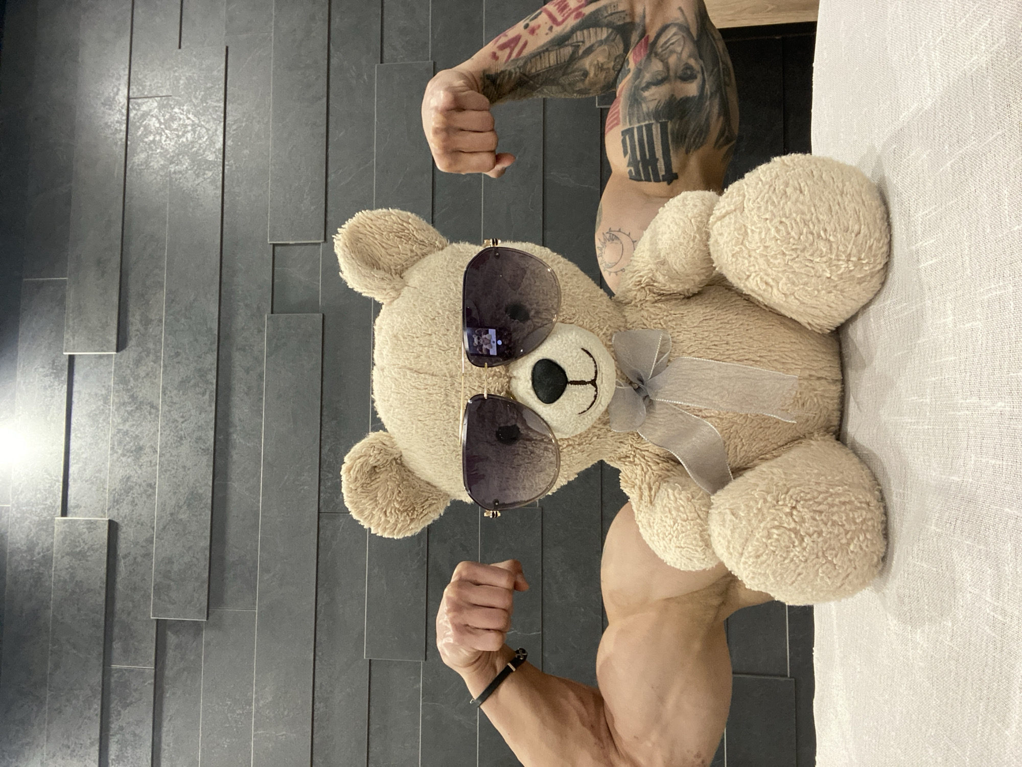 A teddy bear wearing sunglasses is positioned between two muscular arms making fists. The right arm is covered in tattoos. The background includes a textured gray wall on the left and a white textured surface on the right. There is no indication this photo was taken in Pleven, Bulgaria.