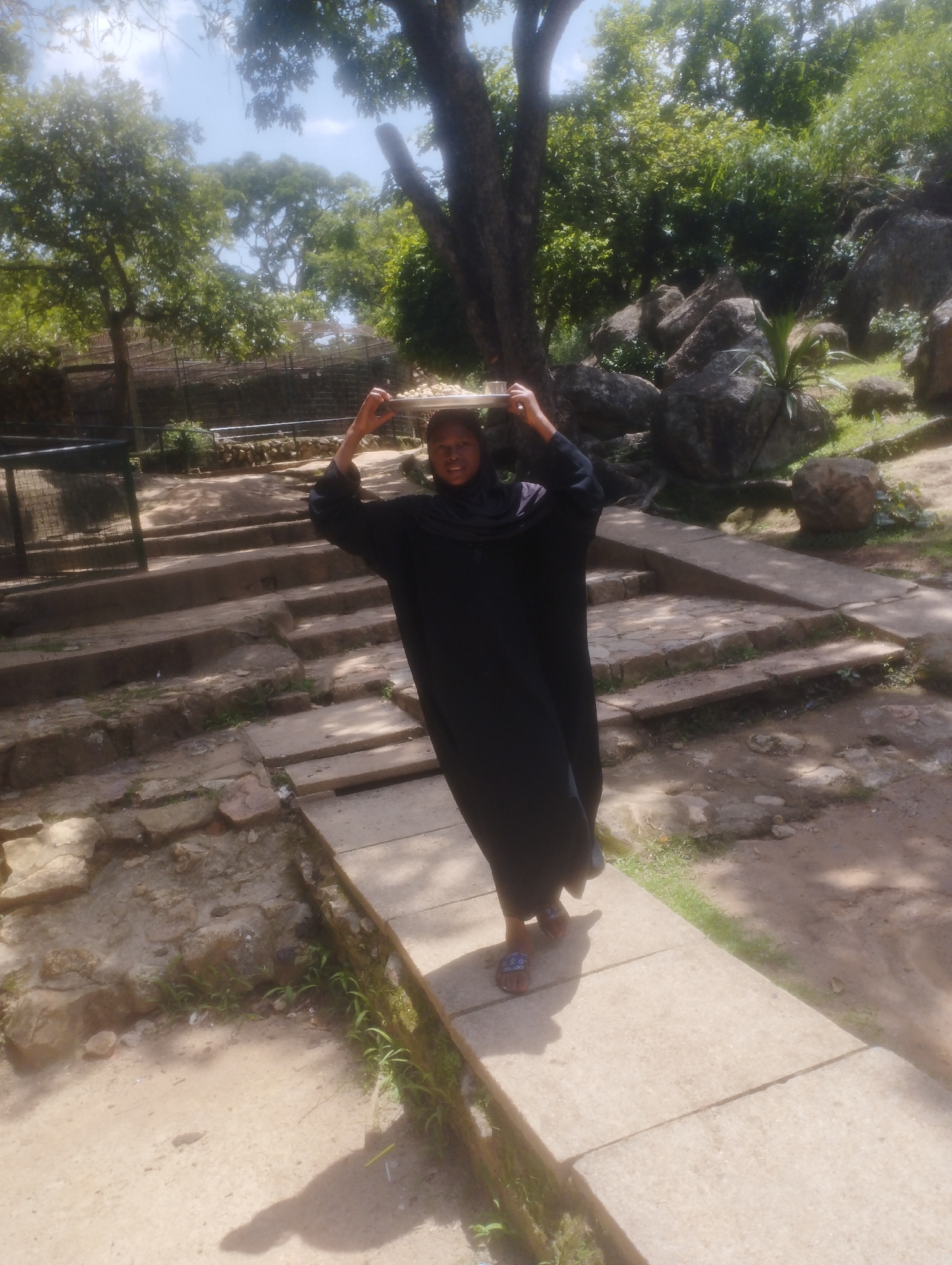 In this outdoor photograph taken in Jos, Nigeria, a young woman is depicted carrying a tray of food on her head. She is dressed in a long, dark abaya and a headscarf, with visible sandals on her feet. The woman is walking on a paved pathway, which is bordered by stone steps and lush greenery. The background features large rocks and trees, suggesting a natural or park-like setting.
The lighting indicates it is daytime, likely with bright sunlight, given the strong shadows cast on the ground. The weather appears to be pleasant, with clear skies visible through the tree canopy. The overall atmosphere is one of everyday activity in a scenic, possibly somewhat rustic, environment. There is no visible text in the image.