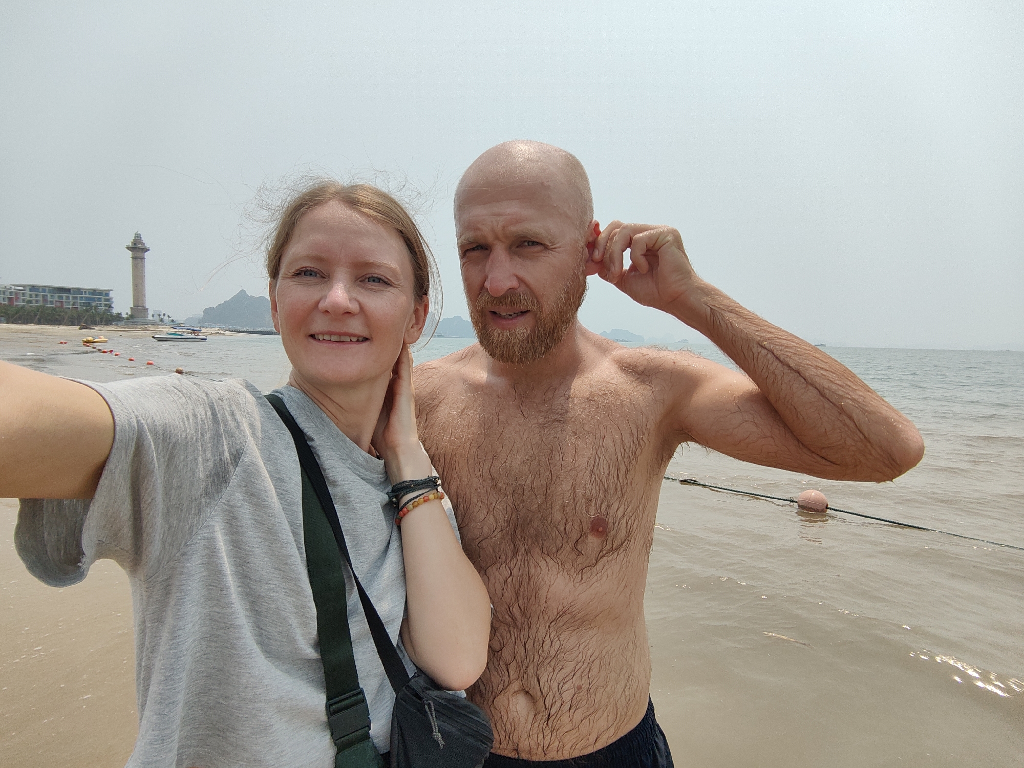 A selfie of two fair-skinned adults on a beach in Bai Chay Ward, Vietnam. The woman is on the left wearing a gray t-shirt and a dark shoulder bag. She is smiling. The man is on the right, shirtless, with a beard and a bald head. His right hand is near his ear. They are standing in shallow water. In the background, a building and a tower are visible, along with boats. The sky is overcast. A rope with floats is visible in the water.