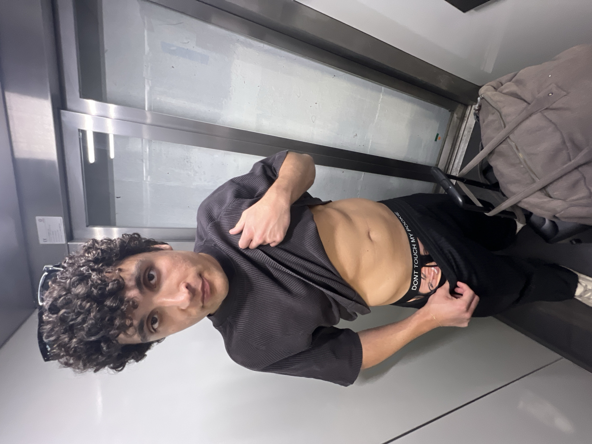 The image captures a young man with dark, curly hair, appearing to be in his late teens or early twenties, looking directly at the camera with a neutral expression. He is wearing a dark, ribbed, short-sleeved shirt, which he has pulled up with his left hand to expose his bare abdomen and navel. With his right hand, he is pulling down the waistband of his black underwear or shorts. A pair of sunglasses is tucked into his hair.
The background shows a modern interior space, characterized by metallic frames and large glass panels, suggesting a public transport vehicle like a train or bus, or a building with extensive windows. In the upper right corner, a large, soft, light brown or grey bag or backpack is visible, resting on what appears to be a seat or bench. A white rectangular label is affixed to a grey wall on the left side of the frame. The floor is a light grey.
The lighting is bright and even, indicating an indoor setting during daytime. The man's pose suggests he is actively showing his physique. On the waistband of his lower garment, the text "DONT TOUCH MY POCKET" is clearly visible, with "ROOKIE" possibly seen below it. The image is set in Milan, Italy.