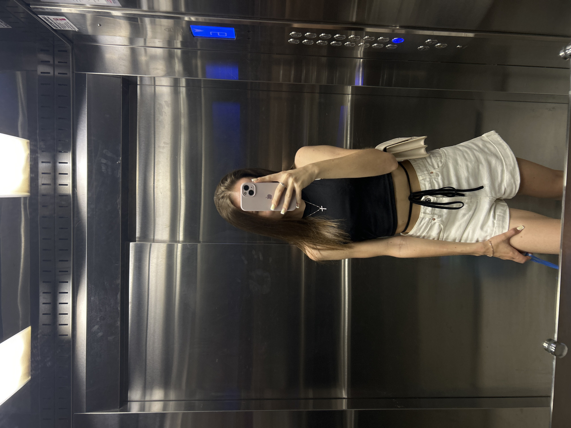 A person is seen taking a mirror selfie inside an elevator. The individual, appearing to be a woman, is positioned in the center of the frame, holding a light pink/purple iPhone with an Apple logo visible on its back, obscuring her face. She is wearing a black crop top, white shorts with black drawstrings, a silver cross necklace, a ring on her left hand, and a bracelet on her right wrist. Her long, brown hair is visible. A small, white purse or bag is slung over her shoulder.
The setting is an elevator interior, characterized by brushed stainless steel walls that reflect light, creating vertical streaks. On the upper right, an elevator control panel is visible, featuring numerous circular buttons and a blue digital display showing illuminated symbols that resemble 'Π0' or a similar floor indicator. To the left, there are vertical strips with horizontal slits, possibly for ventilation or aesthetics. In the bottom right, a blue strap or cord is visible near the person's leg. The lighting within the elevator appears bright, casting reflections on the metallic surfaces. Some faint, unreadable text is visible on warning or instruction signs in the upper left corner.