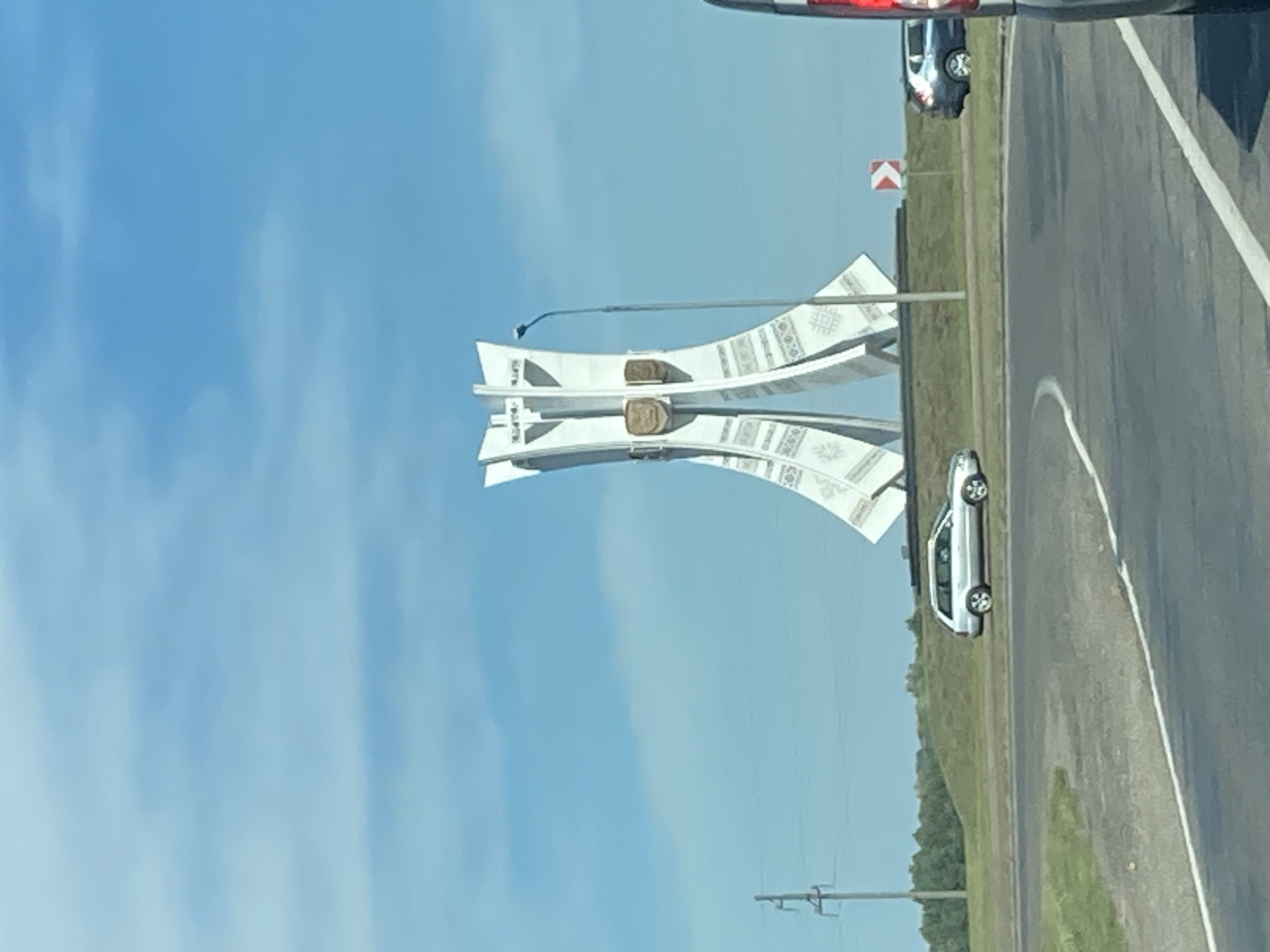 A white, abstract monument stands tall against a bright blue sky with sparse white clouds. The monument has a layered, petal-like structure, decorated with intricate patterns and two crests on its front. A streetlamp stands to its right. In front of the monument is a circular road or roundabout. A silver station wagon drives on the roundabout, and a dark-colored SUV is parked or driving on the right side of the roundabout. The background features a grassy hill lined with trees. On the left side of the image, a utility pole with wires is visible. The scene is captured from a low angle, likely from a vehicle, as evidenced by the road in the foreground and the partial view of another vehicle on the far right. This monument is likely the monument to the Defenders of the Motherland in Brest Fortress, Brest, Belarus.