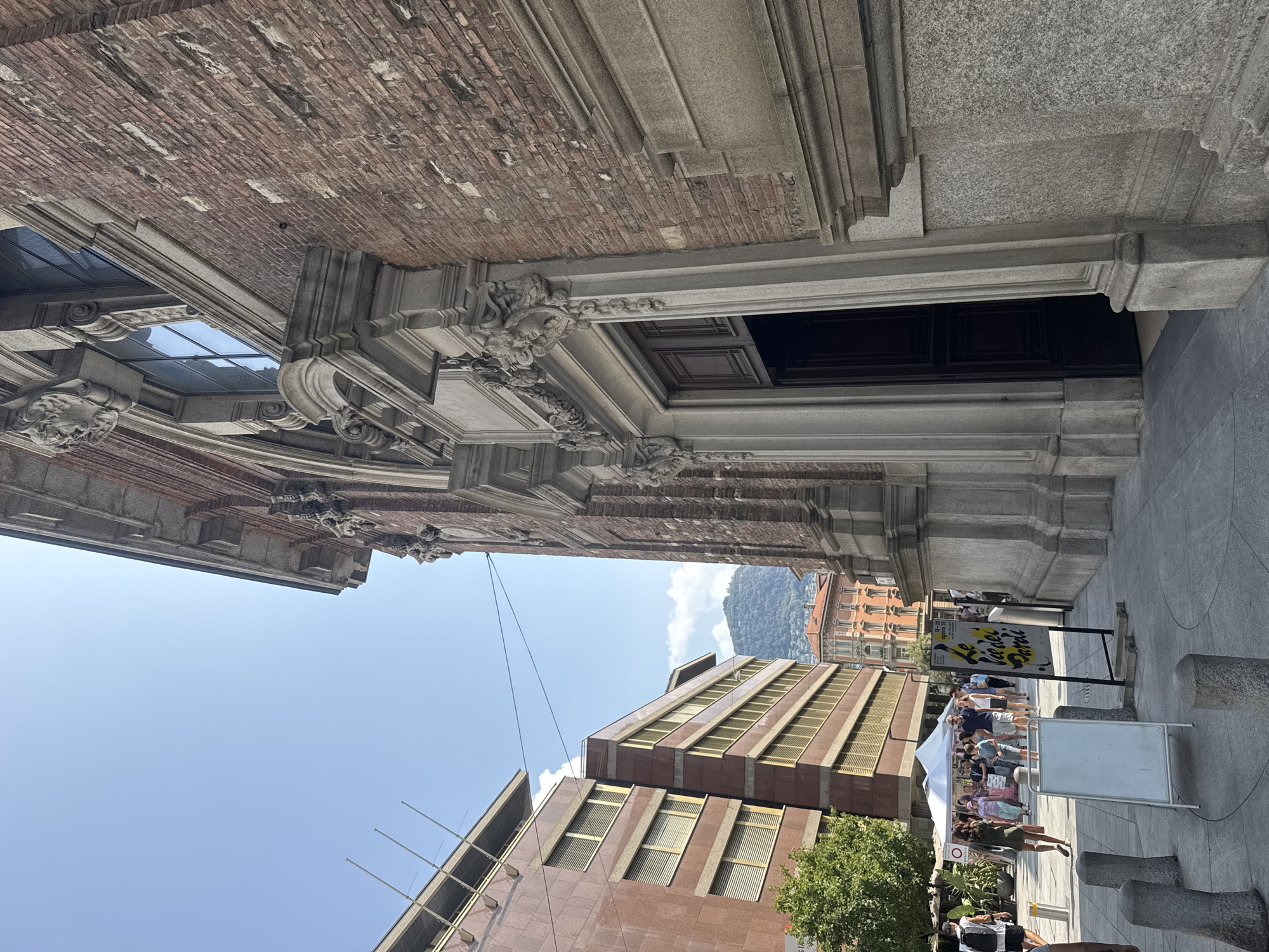 This is an eye-level, outdoor, full-shot photograph taken on a clear, sunny day. The image depicts a street scene in Lugano, Switzerland, specifically in front of the LAC Lugano Arte e Cultura.
On the right side of the frame, a large, ornate brick building dominates the foreground. It features a grand, dark wooden double door entrance, framed by classical architectural elements like columns and decorative carvings of faces and foliage. The brickwork shows signs of age, with some variations in color and texture. A portion of the building's stone base is also visible.
To the left of the brick building, a sidewalk leads towards a street with modern architecture. Two tall, multi-story buildings with reddish-brown facades and horizontal bands of windows are visible. The buildings have a contemporary design, contrasting with the historic structure on the right. In the background, a green, tree-covered mountain can be seen under a clear blue sky.
The street area in front of the buildings is bustling with activity. Several people are walking and gathered. A white tent, possibly for an outdoor market or event, is set up on the left side. There are also signs and whiteboards placed on the sidewalk. One sign, in yellow and black, has text that appears to be in Italian. The ground is paved with gray stones.
The overall impression is a lively urban setting that blends historical and modern architecture, under a bright sky.