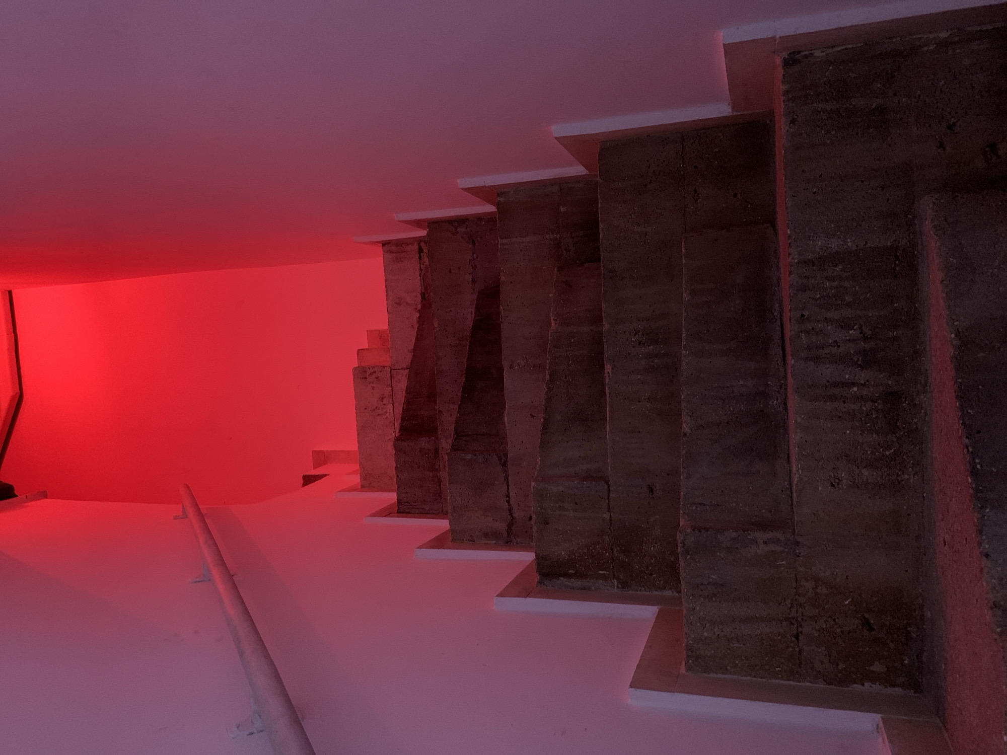 An indoor concrete staircase ascends upwards, illuminated predominantly by an intense red light source originating from above or at the next landing. The stairs are constructed from dark, textured concrete, exhibiting signs of wear and surface imperfections. On the left-hand side, a light-colored metal handrail is affixed to the wall with multiple brackets. The walls, painted a light hue (likely white or off-white), reflect the strong red illumination, appearing in shades of pink and red. The upper section of the staircase and the area it leads to are entirely enveloped in this vibrant red glow. No individuals or specific actions are present in the scene. The precise location within the city of Tel-Aviv, Israel, is not discernible from the provided image.