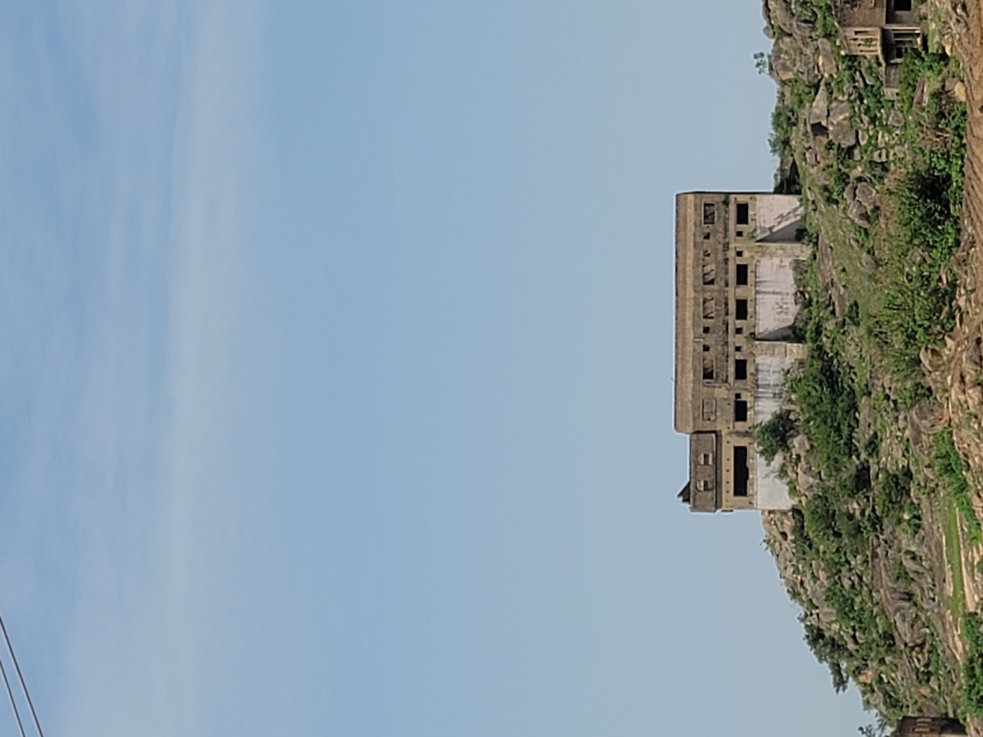 The image shows a large, dilapidated building located on a rocky, vegetated hill in Jos, Nigeria. The building is multi-storied, made of a grey concrete or stone material, and has numerous rectangular window openings. The windows are without glass or frames, suggesting the building is abandoned or in disrepair. The structure is partially obscured by foliage growing on the hillside. The sky is clear and light blue, with a few faint clouds visible. A power line is running along the left side. The image is oriented sideways.