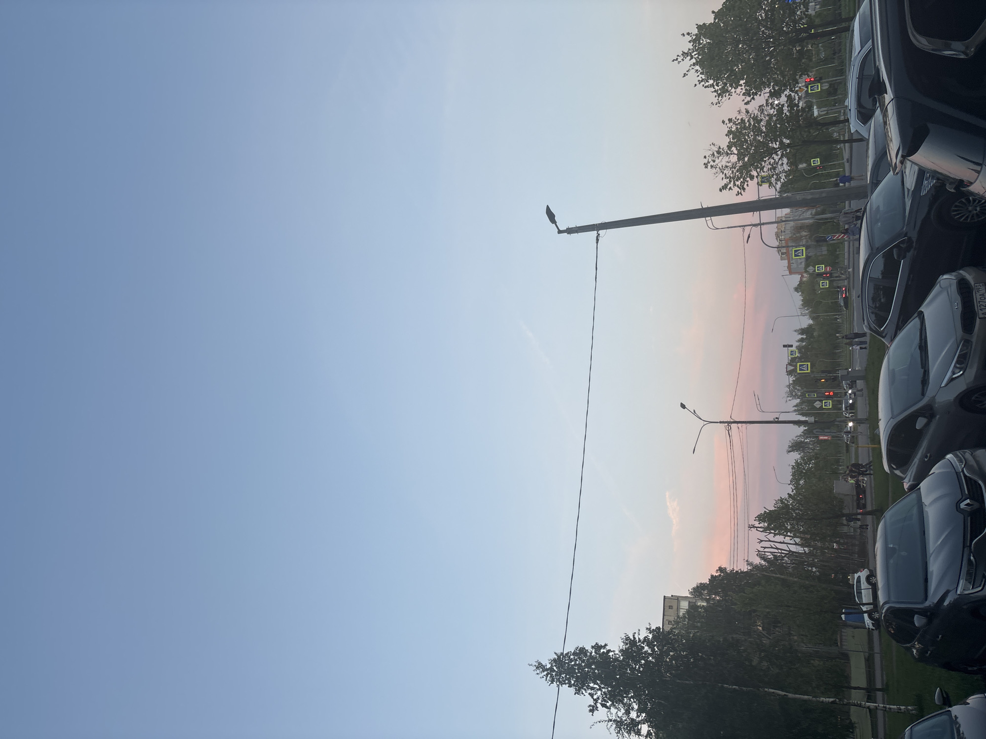 The image captures a cityscape at dusk or dawn. The sky transitions from a light blue to a soft pinkish hue near the horizon. Utility poles and overhead wires stretch across the sky. A row of parked cars lines the right side of the frame. Trees with sparse foliage are visible in the background. Street signs and traffic lights can be discerned in the distance. The location appears to be a residential or commercial area in an unidentified city.