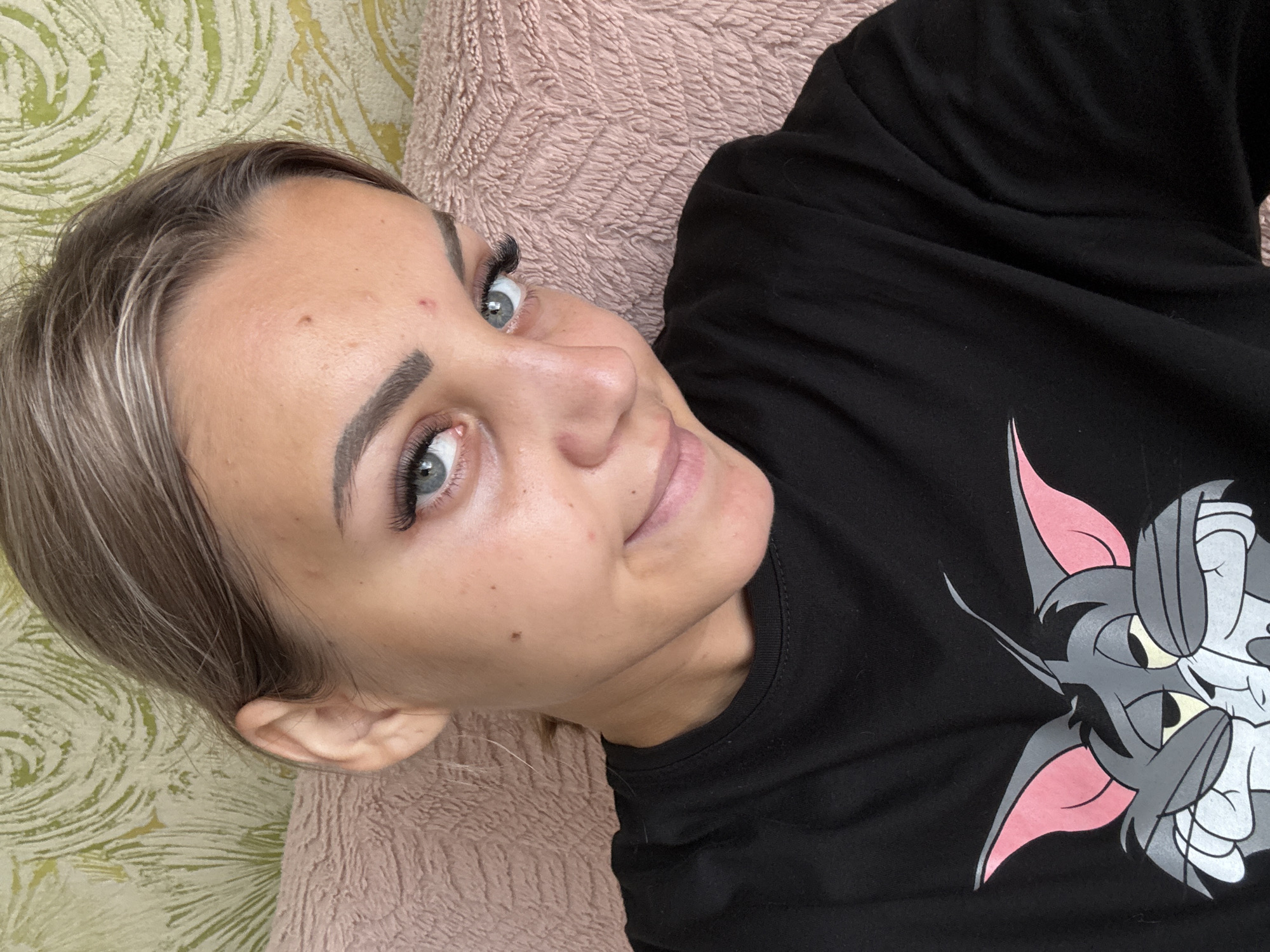 A woman is taking a selfie, likely in her home in Krasnogorskoye, Russia. She has light brown hair styled away from her face, blue eyes, and light skin with some blemishes. She is wearing a black t-shirt with a cartoon image of Tom from Tom and Jerry on it. The background appears to be a patterned wallpaper and a pink textured pillow. The lighting suggests it is daytime, and the focus is on her face and upper body.