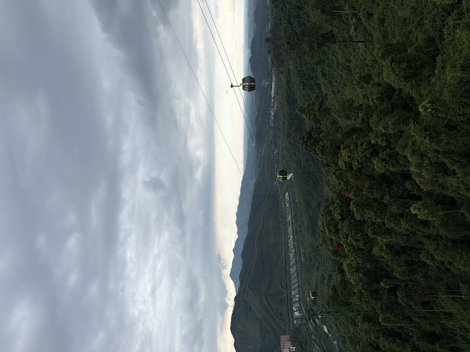 The image shows a view from the Bà Nà Hills cable car in Đà Nẵng, Vietnam. The scene captures several cable cars suspended on wires, positioned over a landscape that includes a combination of mountains, dense green forests, and a visible city area in the background. The sky is cloudy and overcast, covering a significant portion of the frame.