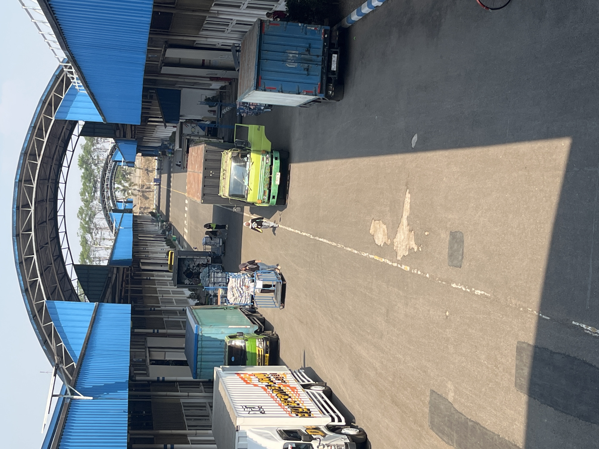 The image depicts an outdoor loading and unloading area, likely at a logistics hub or industrial facility, under a covered walkway. The structure features a prominent blue corrugated metal roof supported by a white metal framework, with additional blue metal siding forming the walls of the covered area. The walkway extends into the distance, with multiple truck bays or sections visible. The ground is paved asphalt, with some patches and markings, including a dashed white line running down the center and a solid yellow line on the left side.
Several vehicles are present in the scene. On the left, a white truck with a large graphic on its side is parked. To its right, a light green truck is positioned next to a large teal shipping container. Further down the lane, a blue shipping container truck is parked on the right.
There are a few individuals visible. Two people, possibly workers, are standing near a blue cart loaded with sacks, which is positioned between the white truck and the light green truck. Another person in a hijab is walking away from the camera in the middle of the lane. In the distance, other figures appear to be working or moving around.
The environment suggests a sunny day, indicated by the strong shadows cast by the structures and vehicles. The background shows trees and possibly other industrial buildings. The overall scene is one of commercial activity and logistics operations. The location is specified as Pelang, Indonesia.