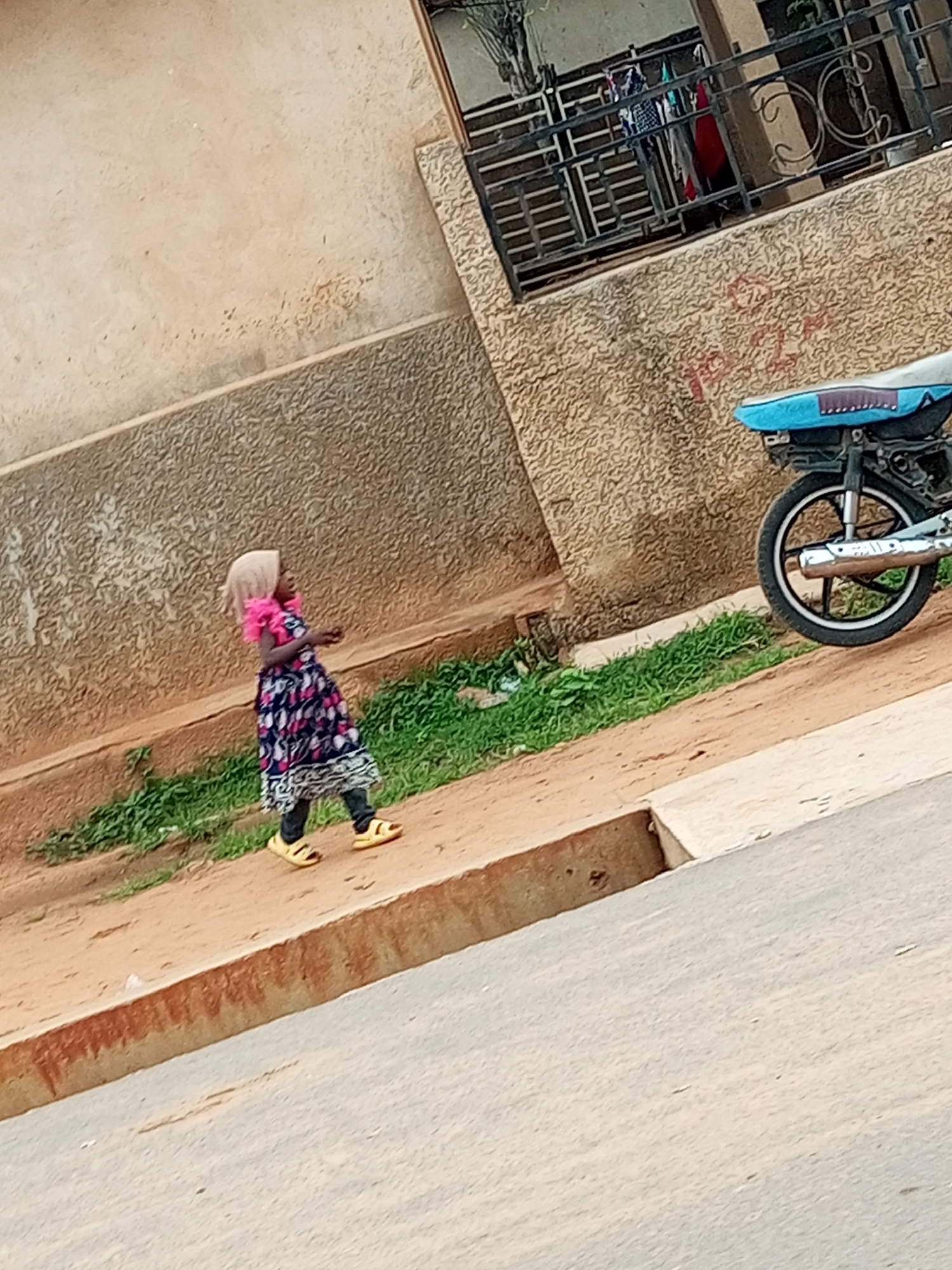 A young child wearing a light-colored headscarf, a pink frilly collar, and a dark dress with a floral pattern walks on a sandy path beside a low wall. The child is wearing yellow sandals. Behind the child, there is a patch of green grass. Further back, a motorcycle with a blue seat is parked next to a textured, light-brown wall. The wall has some red graffiti on it. Above the wall, a metal railing with decorative swirls can be seen, with laundry hanging behind it. The ground in the foreground is a light gray asphalt. The scene appears to be on a street in Tempure, Nigeria, with a building visible in the background.