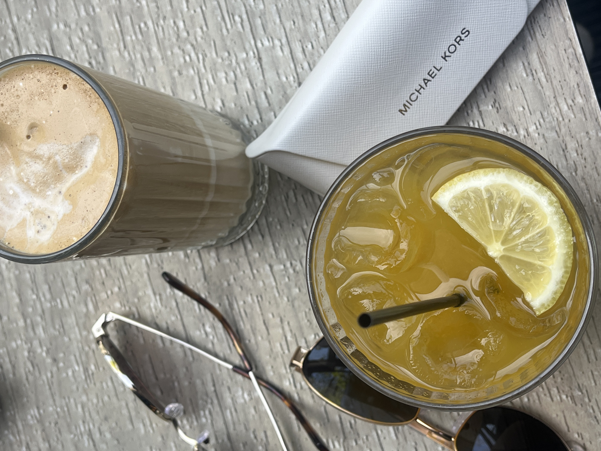 This overhead shot depicts a casual outdoor scene, likely at a cafe or restaurant in Plovdiv, Bulgaria, during daytime. Two beverages and personal accessories are arranged on a light-colored, textured table, possibly made of wood or stone.
On the left, a tall glass holds a frothy, light brown drink, resembling an iced coffee or latte, with visible foam and bubbles. Next to it, a white, subtly textured rectangular case is positioned, clearly displaying the brand name "MICHAEL KORS" in black capital letters.
On the right side of the frame, another glass contains a vibrant yellow, iced beverage, garnished with a fresh lemon slice and equipped with a dark straw, suggesting it could be lemonade or iced tea. Two pairs of sunglasses are also visible on the table: one with a slender, metallic frame (possibly silver or chrome) and another with a thicker, gold-toned frame and dark lenses.
The presence of iced drinks and sunglasses indicates warm or sunny weather, suggesting a leisurely moment spent outdoors. The scene evokes a feeling of relaxation and refreshment.