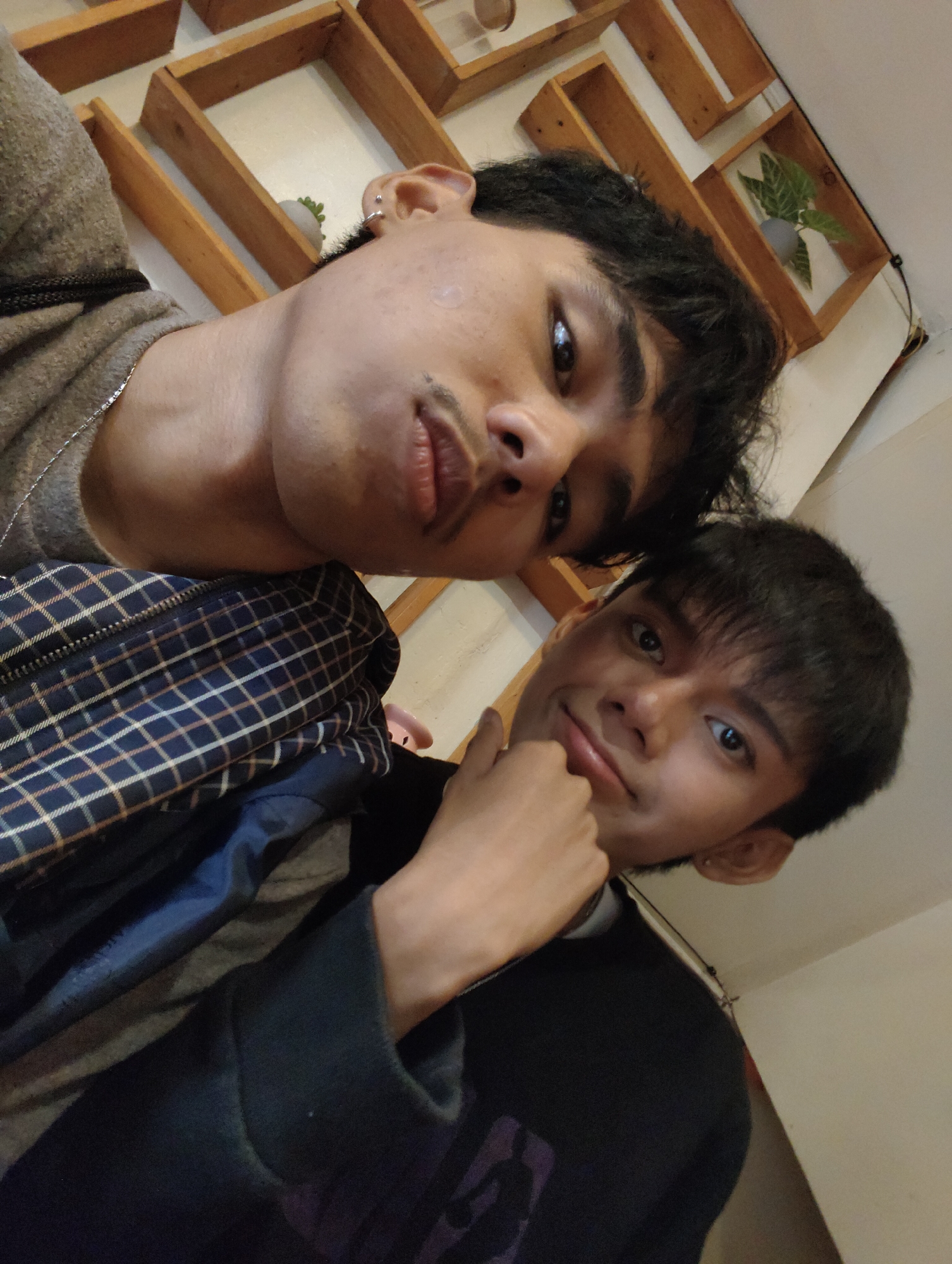 Two individuals are positioned in a selfie with their faces close together. The person in the foreground has dark hair and is looking at the camera with a slight smile, resting their chin on their hand. They are wearing a dark long-sleeved shirt. The second individual, slightly behind and to the left, has dark, wavy hair and is also looking towards the camera, with their face angled. They are wearing a grey sweater and a silver necklace. A checkered pattern of a jacket or bag is visible between the two individuals. The background consists of a white wall with wooden shelves, one of which contains a small potted plant. The lighting suggests an indoor environment. The image is oriented in a way that suggests it was taken from a low angle. Without further information, it is not possible to determine the specific location within Davao City, Philippines.