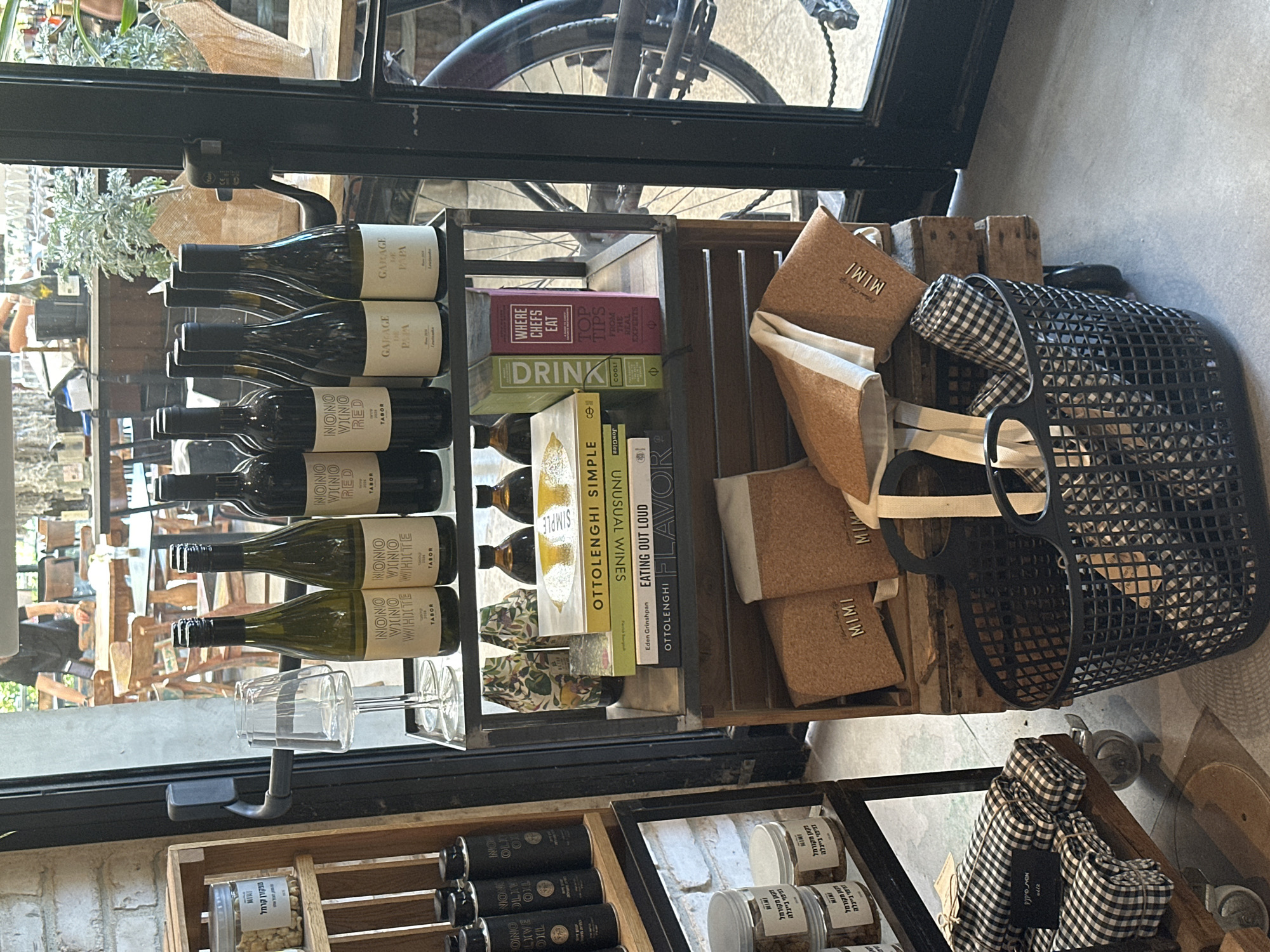The image presents an interior retail display within what appears to be a cafe or specialty food and wine shop in Petah Tikva, Israel. The scene is illuminated by natural light entering through a large glass door on the right, which offers a view of an outdoor or glass-enclosed dining area with wooden tables and chairs, and a partially visible bicycle.
On the left, a multi-tiered wooden shelving unit is stocked with various packaged food items. The top and bottom shelves hold clear glass jars, each bearing the "MIMI" brand and Hebrew labels indicating "מקורמל" (caramelized), "פיקנטית" (pikantit), and "ג'ינג'ר יפני" (Japanese ginger) for dried goods, likely spices or nuts. The middle shelf displays several black cylindrical bottles identified as "NONO OLIVE OIL" with accompanying Hebrew text. Below the shelves, a small wooden trolley with wheels carries stacks of neatly folded black and white checkered fabric items, marked with a small black tag reading "אסי אקססוריז" (Assi Accessories).
In the central foreground, a dark metal shelving unit serves as the primary display. The top shelf features two empty clear wine glasses alongside eight wine bottles. From left to right, these include two green bottles labeled "NONO VINO WHITE TABOR," one dark bottle labeled "NONO VINO RED TABOR," and five dark bottles labeled "GARAGE DE PAPA Laurieska." The presence of "TABOR" indicates an Israeli winery. The middle shelf is organized with five horizontally stacked books on culinary and wine themes, specifically "OTTOLENGHI SIMPLE," "UNUSUAL WINES" (with Hebrew "יינות לא שגרתיים"), "EATING OUT LOUD," and "OTTOLENGHI FLAVOR." Two additional books, "DRINK" and "WHERE CHEFS EAT," are stacked vertically. This shelf also holds several small, dark amber bottles and two decorative bottles with botanical patterns. The bottom section of the display showcases a large, black, grid-patterned plastic market basket containing more black and white checkered fabric items, situated on a wooden crate. Adjacent to it are several cork-colored pouches or clutches, each embossed with the "MIMI" brand, some featuring white fabric straps.
The floor is a light grey concrete. The visible wall on the left is of a light-colored brick or stucco texture. A person in a dark shirt is partially visible through the glass in the background.