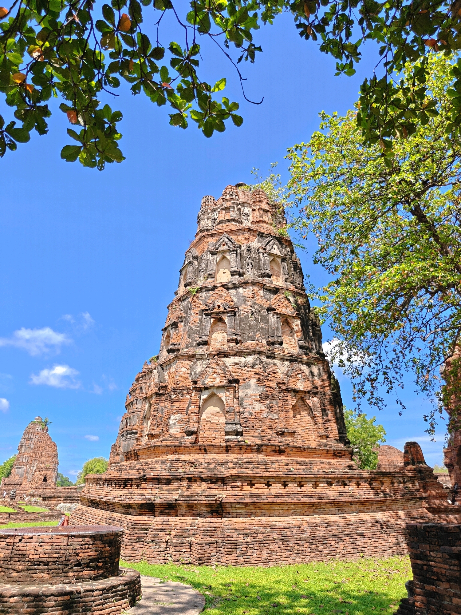 The image depicts the ancient ruins of Wat Mahathat in Phra Nakhon Si Ayutthaya City Municipality, Thailand. The central subject is a large, weathered brick stupa, its tiered structure and intricate detailing indicative of Ayutthaya-era architecture. Surrounding it are other brick structures, some in advanced states of ruin, creating a sense of historical depth. Lush green grass covers the ground, and the clear blue sky is partially obscured by the branches of leafy trees, casting dappled shadows on the ruins. The scene is bathed in bright sunlight, highlighting the texture of the ancient bricks and the vibrant green of the vegetation. There are no people visible in the image.