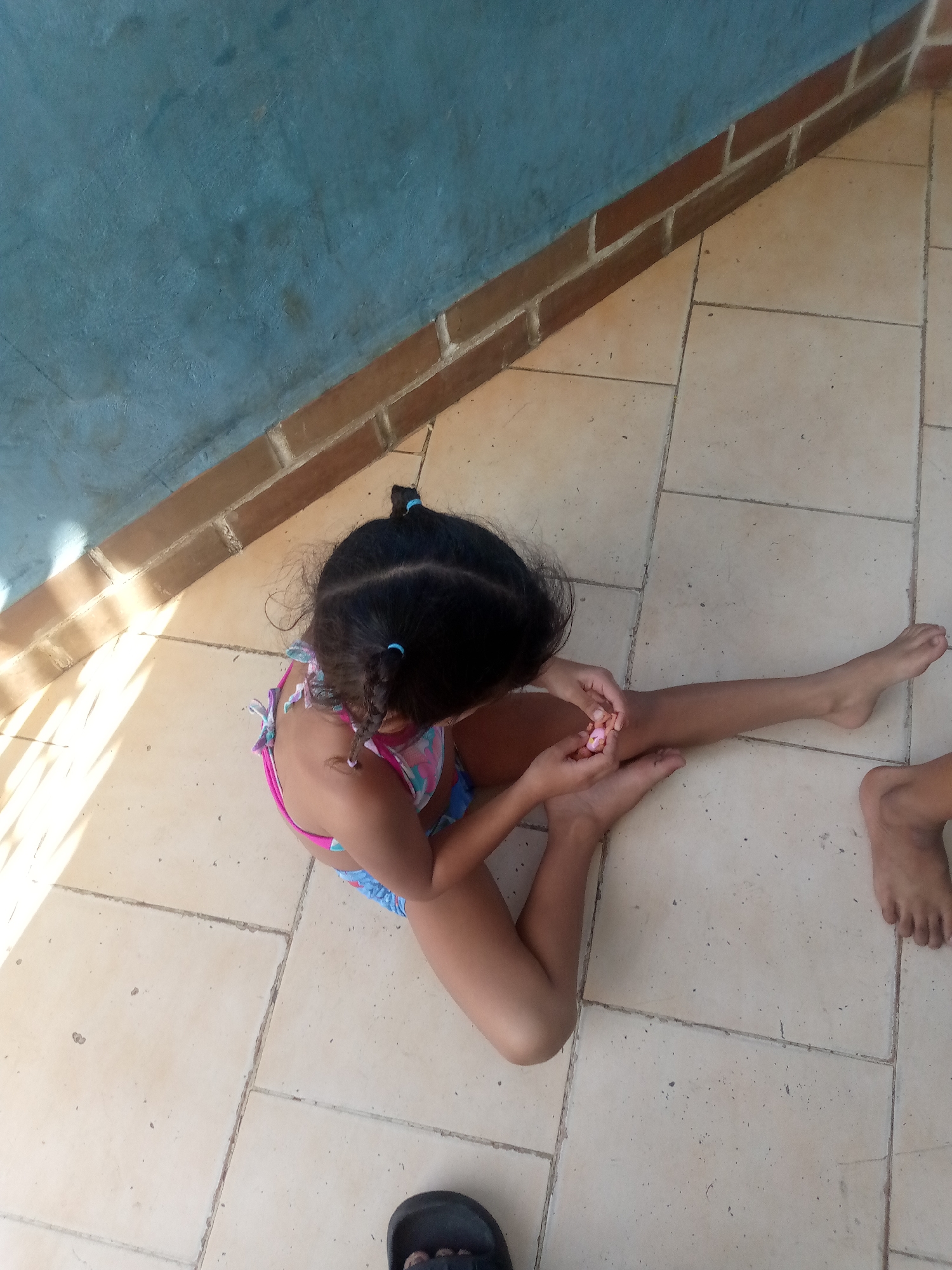 A young girl, seen from a high angle, sits on a tiled floor next to a light blue wall. She is wearing a colorful bikini. Her dark hair is pulled back into two braids with blue ties. The girl is holding a small, pink object in her hands and appears to be examining it. Sunlight streams in from the left, casting striped shadows across the floor. Another person's bare feet are visible on the right side of the frame, and a black sandal is partially visible at the bottom. The scene appears to be outdoors, possibly in a patio or yard, given the tiled floor and the presence of sunlight. There is no visible text in the image.