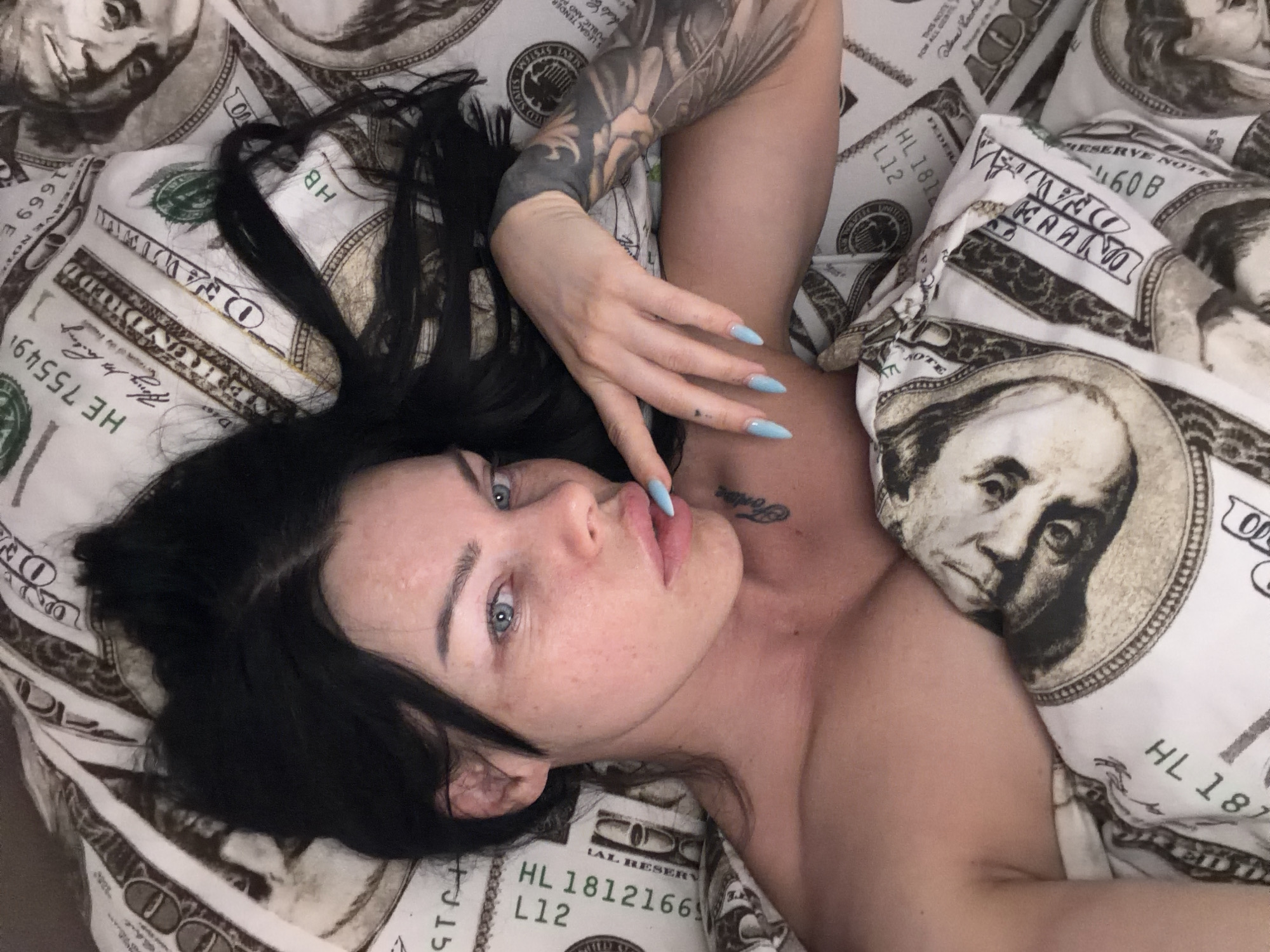 A young woman with dark hair and blue eyes lies on a bed covered in a comforter printed with images of US $100 bills. She is looking up and to her right, with her right hand resting on her face, near her mouth. Her fingernails are painted light blue. She has tattoos on her left arm and a small tattoo on her upper chest. The scene is lit with a soft, natural light. There is no discernible location in the city of Ufa, Russia.