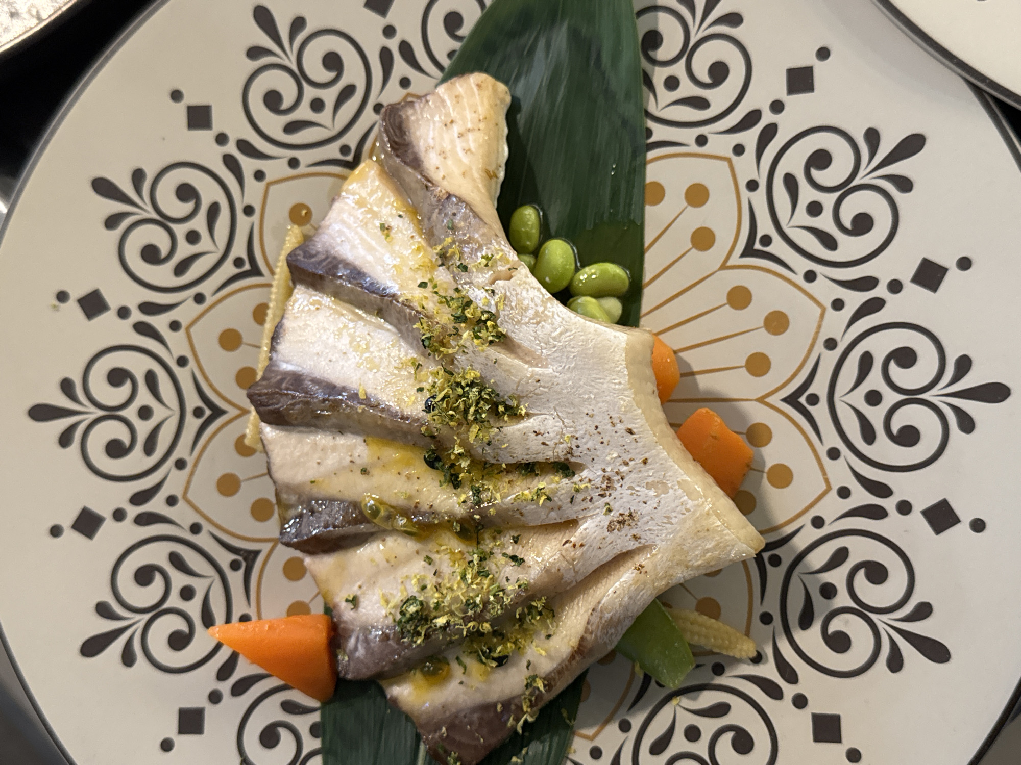 The image displays a close-up, overhead view of a dish of fish served on a decorative plate. The fish appears to be thinly sliced and fanned out, garnished with a green herb and what looks like lemon zest. Beside the fish, there are several edamame beans, a small piece of orange carrot, and a sliver of baby corn, all resting on a large green leaf. The plate itself has a complex, symmetrical pattern in black, gold, and off-white. The lighting suggests an indoor setting, likely a restaurant. There is no indication of the city of Sofia, Bulgaria, or any people present in the image.