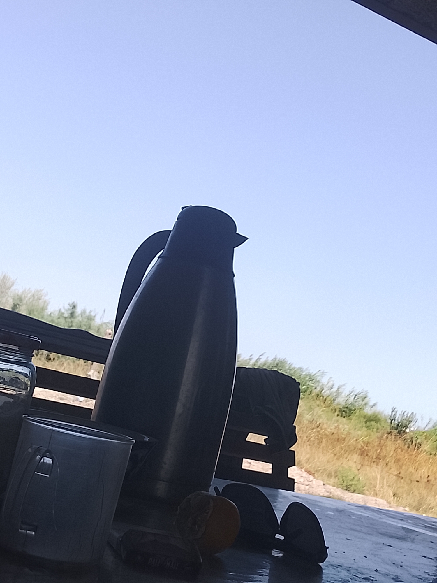 The image depicts an outdoor scene with a clear blue sky. In the foreground, a dark thermos is positioned on a table, with its handle clearly visible. To the left of the thermos, a metallic cup is also present. Further to the left, a glass jar is partially visible. On the table, there are also two dark circular objects and a small orange fruit. In the background, there is vegetation and a wooden structure. The image appears to be taken in Sabir Rakhimov, Uzbekistan, given the context provided.