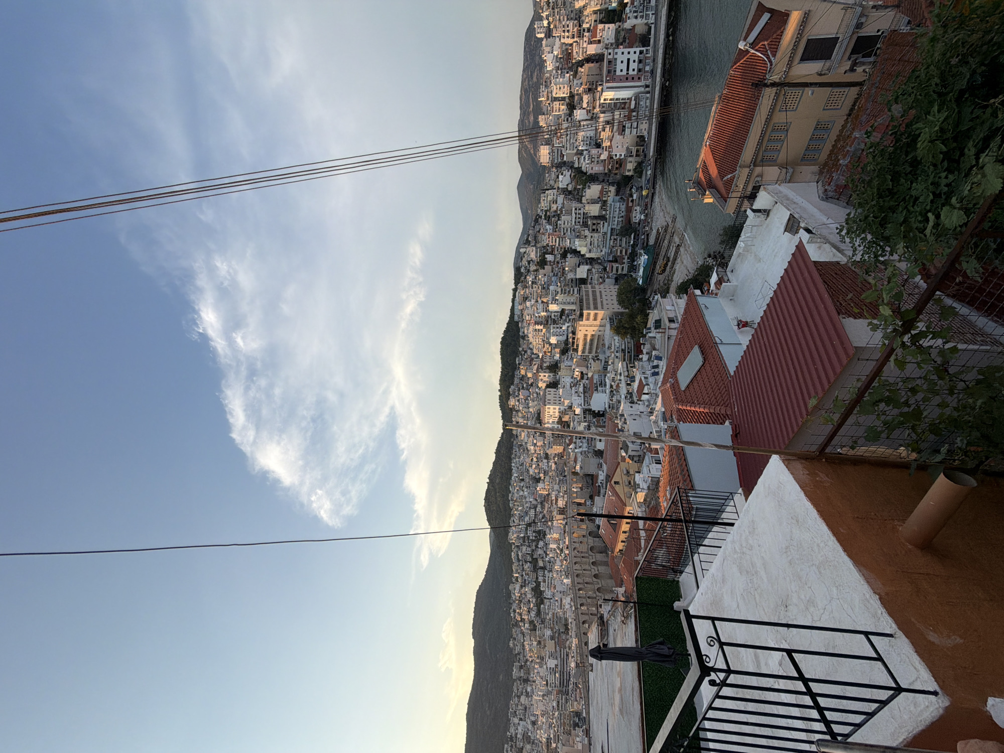 The image captures an elevated view of the city of Kavala, Greece, at dusk. In the foreground, a section of a rooftop is visible with white and orange painted walls, a black metal railing, and some green foliage. A large pipe protrudes from the orange wall. Beyond the rooftop, the city sprawls across a hillside towards the sea. Numerous buildings with red-tiled roofs and white or light-colored facades dominate the urban landscape. A prominent aqueduct, a historical landmark, stretches across a section of the city in the mid-ground. The sea is visible to the right, with a few boats or structures near the shore. The sky is a soft blue with a few wispy clouds, illuminated by the setting sun. Several power lines traverse the frame from top to bottom. The scene suggests a peaceful evening view of a coastal city.
