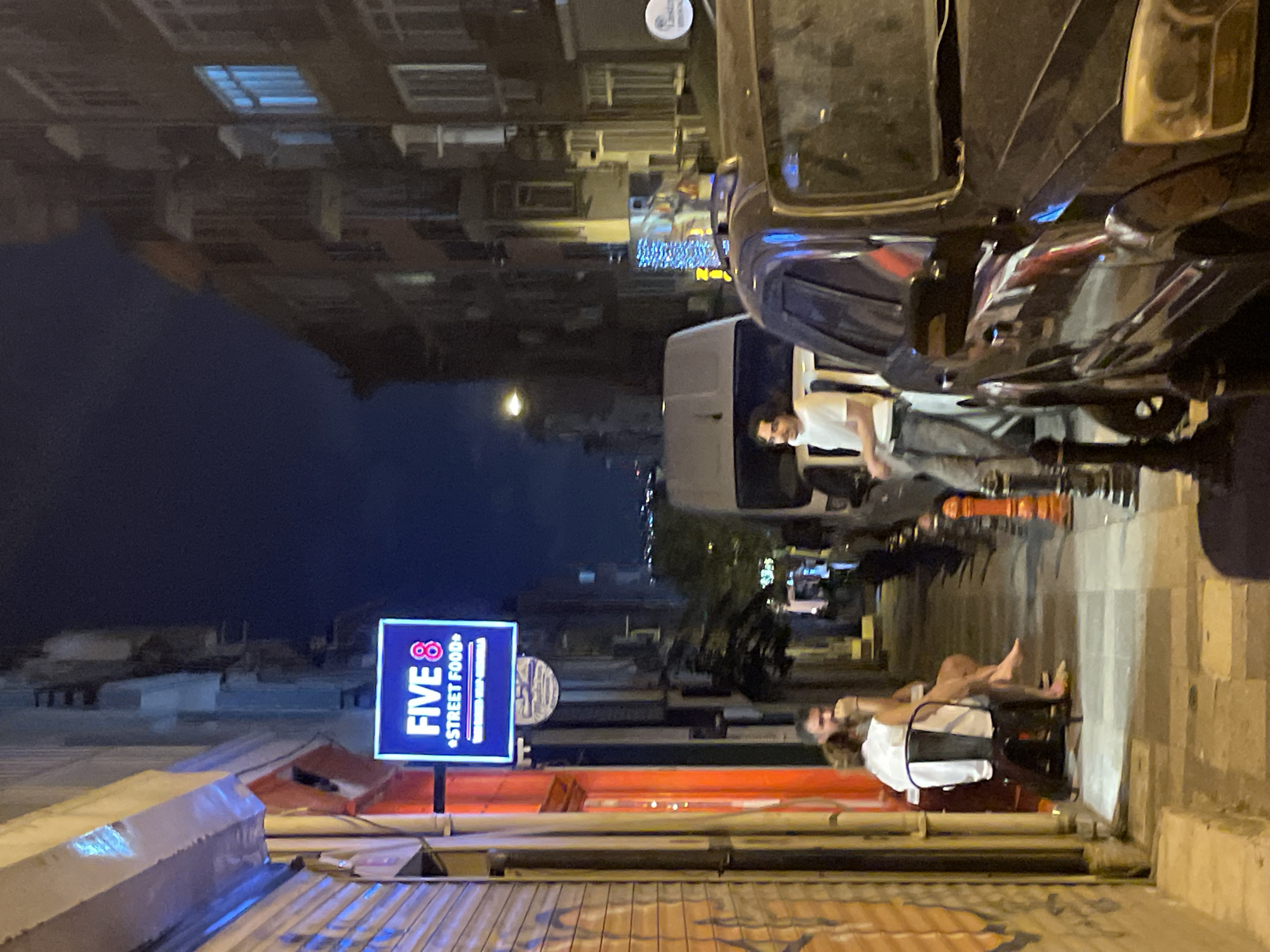 The image depicts a nighttime scene on a street in Kadıköy, Turkey, likely a pedestrian walkway or narrow street. A lit neon sign reading "FIVE 8 STREET FOOD" is prominently displayed on the left side of the frame, above an entrance to a establishment with an orange-tinted facade. Two individuals, a man and a woman, are seated at a small table outside this establishment, facing away from the camera. The man is wearing a white shirt, and the woman is in a white dress, with her legs visible.
Further down the street, a young man with dark, wavy hair, wearing a white t-shirt and grey pants, stands by a white van. His posture suggests he is waiting or observing. In the foreground, the front portion of a dark-colored SUV is visible, obscuring part of the street and the white van. The street is lined with buildings on both sides, their windows illuminated with a mix of warm and cool-toned lights, suggesting residential or commercial spaces. The atmosphere is urban, with a sense of quiet activity on a cool night.